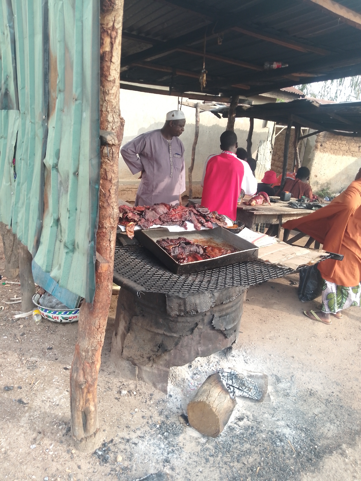 The image depicts an outdoor food stall in Serti, Nigeria, specializing in grilled meat. In the foreground, a large metal tray filled with cooked meat rests on a grill made of metal bars. Below the grill is a large metal drum, possibly used as a charcoal burner. To the left, a corrugated metal sheet structure, painted a faded green, serves as a makeshift wall for the stall, with a rough wooden post supporting it. In the background, several individuals are visible. An older man wearing a white cap and a purple robe stands to the left, facing away from the viewer. Behind him, a person in a red shirt is partially visible. Further back, other people are gathered around tables, suggesting a bustling market environment. The ground is sandy and scattered with ash and small stones, indicating the presence of fire and cooking activities. The overall scene conveys a sense of everyday commerce and food preparation in a Nigerian setting.