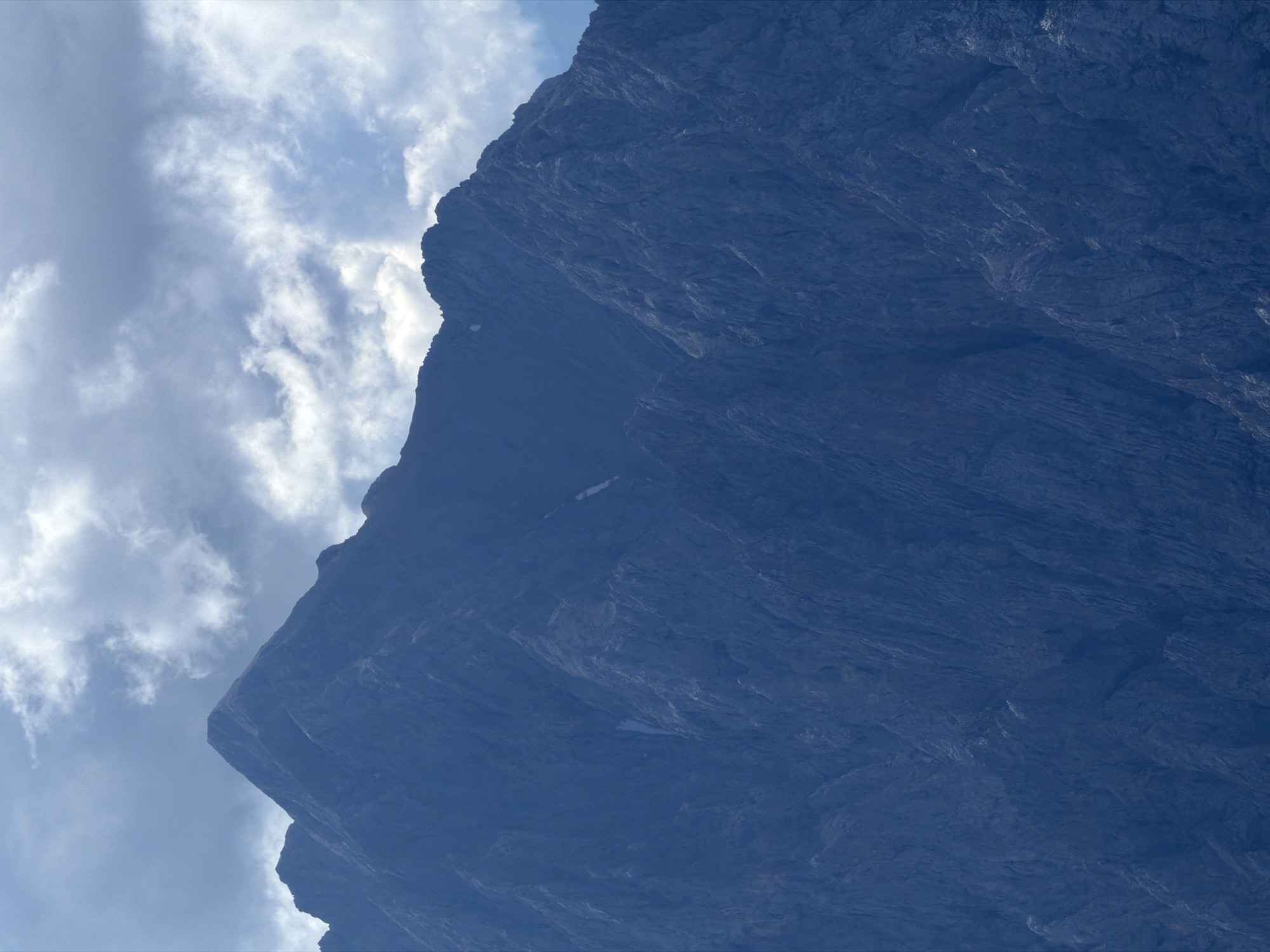 The image shows a close-up view of a dark blue, rocky mountain peak against a cloudy sky. The mountain dominates the frame, with its rugged, textured surface indicating steep inclines and sharp ridges. Patches of snow are visible in some of the crevices and gullies on the mountain face. The sky above is filled with a mix of gray and white clouds, with some areas of bright light suggesting the sun is partially visible. The overall impression is of a vast and imposing natural landscape. This mountain is Zugspitze, the highest peak in Germany, located on the border between Germany and Austria, near Garmisch-Partenkirchen, Germany. There are no people, animals, or man-made objects visible in the image.
