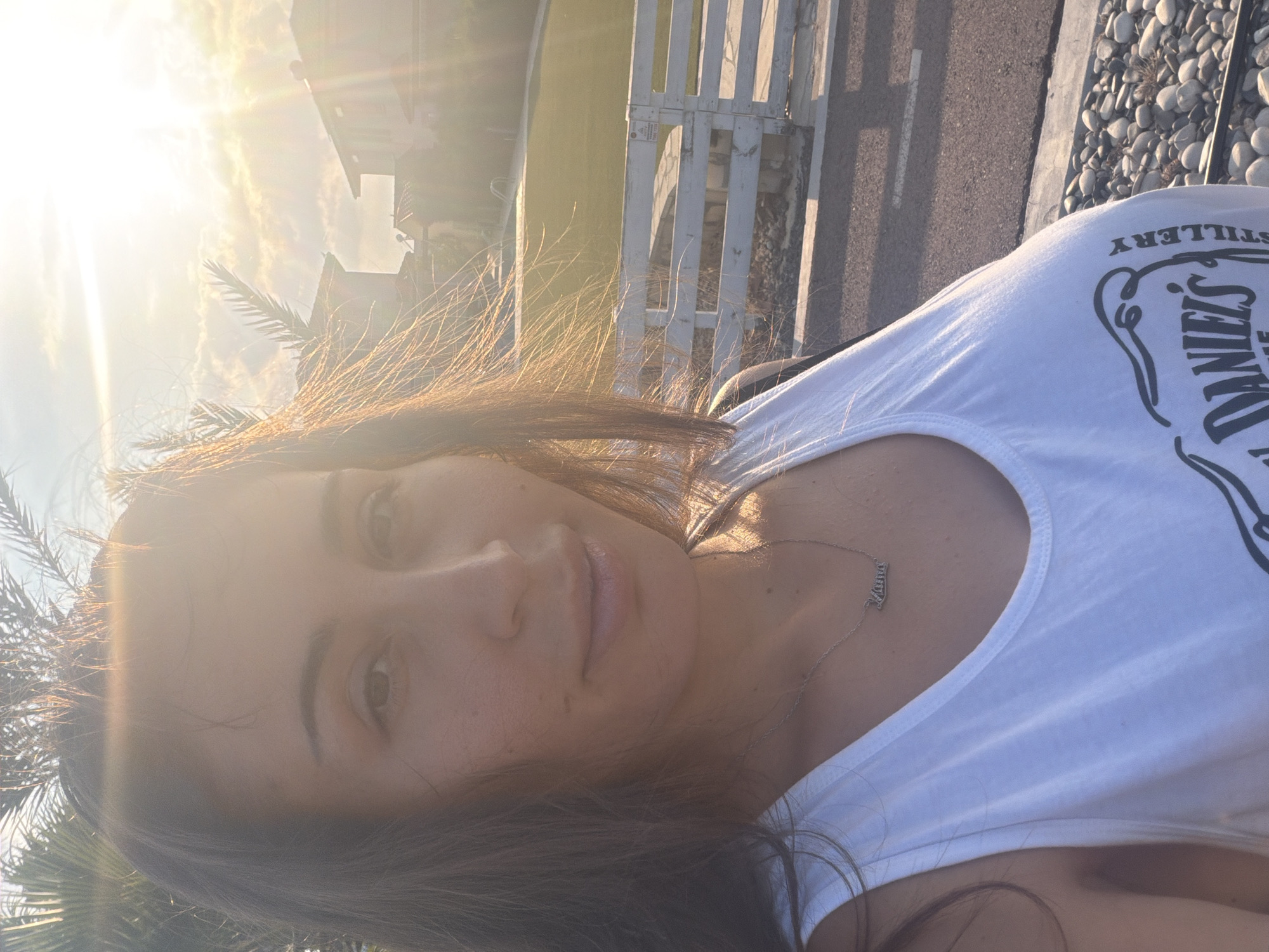 A woman is taking a selfie outdoors in Meneou, Cyprus. She has dark hair and is wearing a white tank top with a necklace. The sun is setting, casting a warm glow on the scene. In the background, there are palm trees, houses, a white fence, and a swimming pool. The image appears to be taken in a residential area.