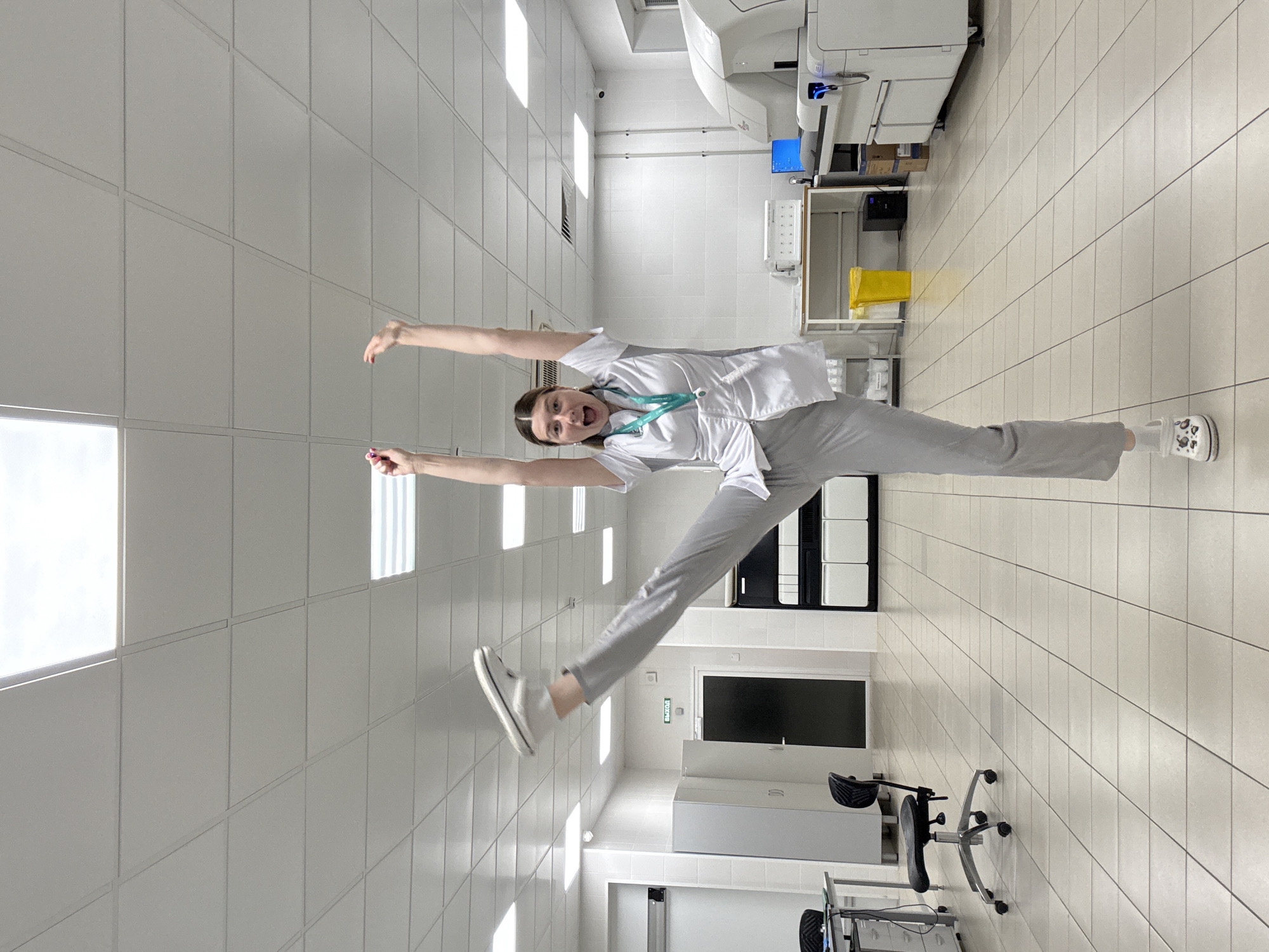 In a brightly lit, tiled interior resembling a laboratory or medical facility, a young woman in a white lab coat and grey scrubs strikes a dynamic pose. Her left leg is extended straight upwards, while her right leg supports her weight. Her arms are raised above her head, and her mouth is open in an expression of excitement or surprise. The room contains various pieces of medical equipment, including a large, white automated analyzer on the right side of the frame, and a desk with a computer monitor and other supplies in the background. Fluorescent lights are embedded in the white drop ceiling. The overall impression is one of playful energy in a professional setting.