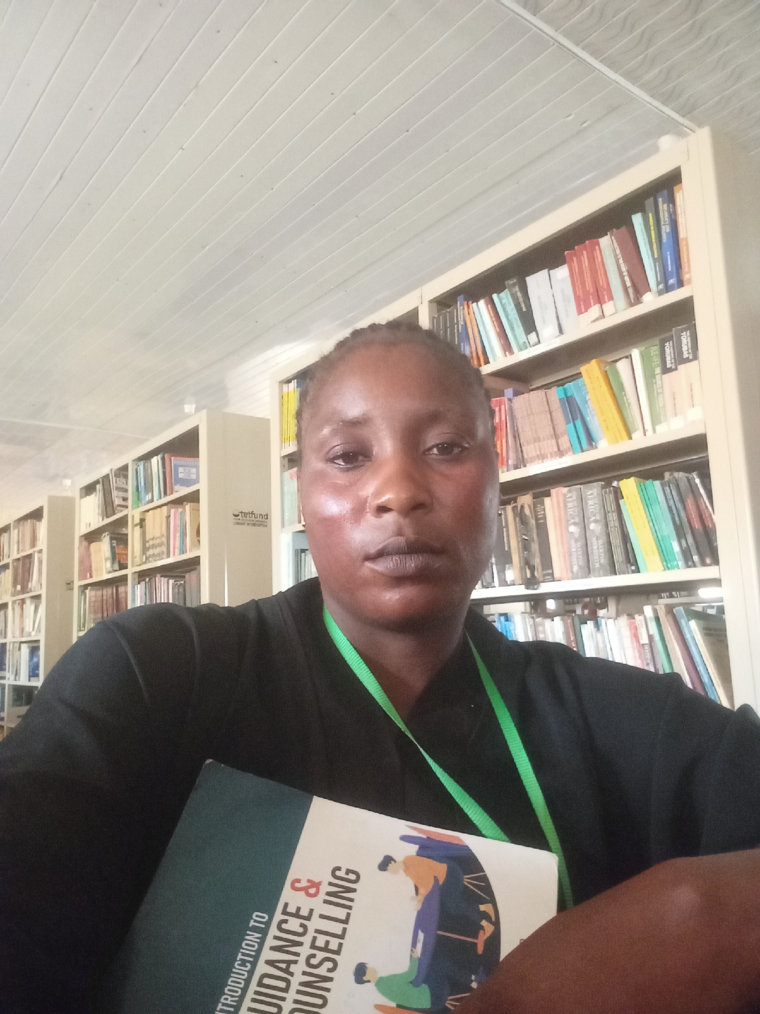The image shows a person holding a book titled "Introduction to Guidance & Counselling" in what appears to be a library in Akwanga, Nigeria. The person is wearing a black top and a green lanyard. Shelves filled with books are visible in the background. The ceiling has a panelled design. The lighting is bright, suggesting an indoor environment.