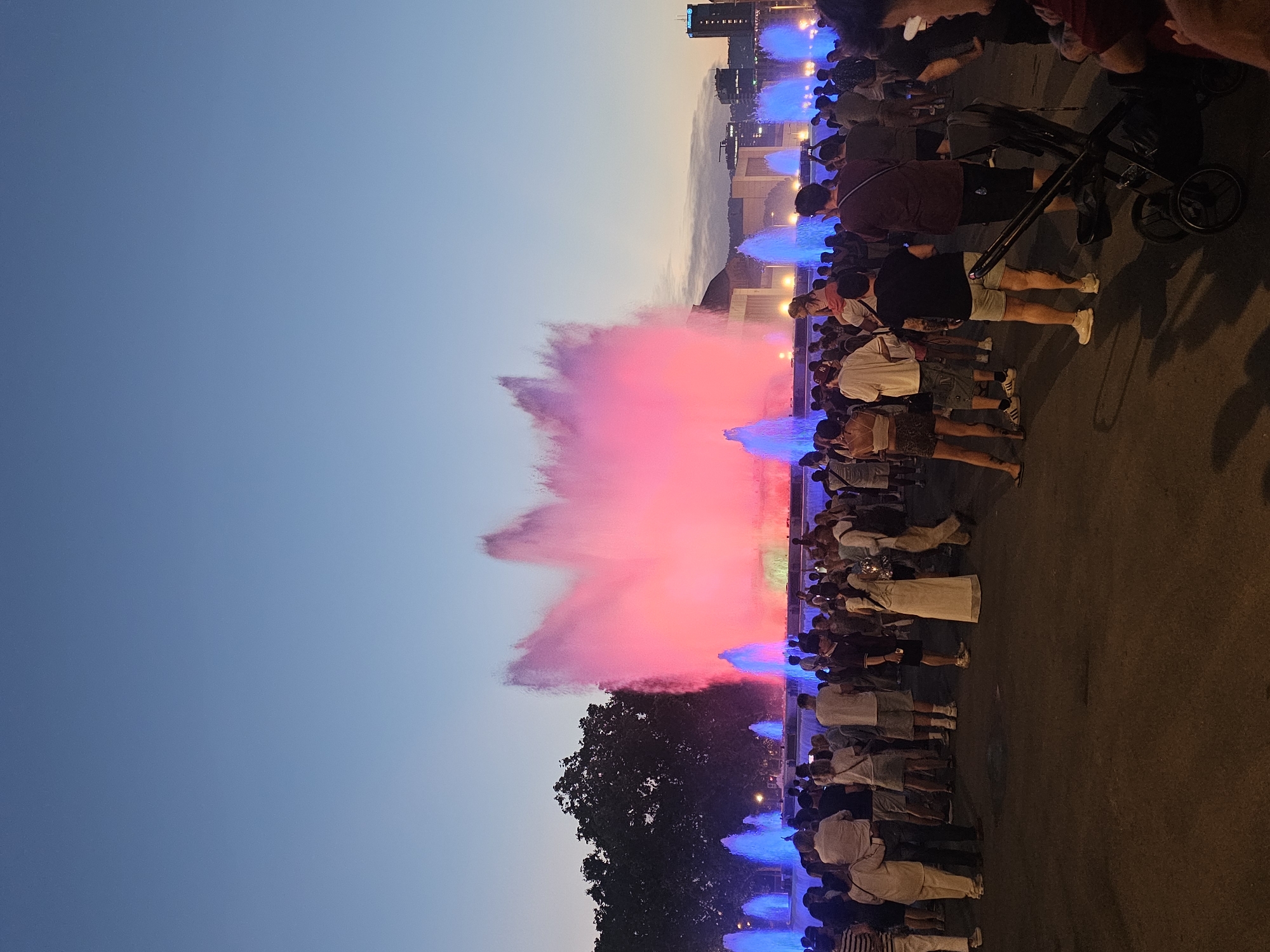The image depicts a crowd of people gathered to watch a colorful fountain show at dusk. The sky transitions from a clear, pale blue to a soft gradient of twilight hues. The fountains are illuminated with vibrant pink and blue lights, creating a dramatic display of illuminated mist and water jets. The crowd appears engaged and appreciative of the spectacle, with many holding up phones to capture the moment. Some individuals are standing, while others are seated on the ground or in strollers. The scene suggests a public event or performance, possibly a tourist attraction. In the background, modern buildings are visible, hinting at an urban setting. The overall atmosphere is one of shared enjoyment and wonder.