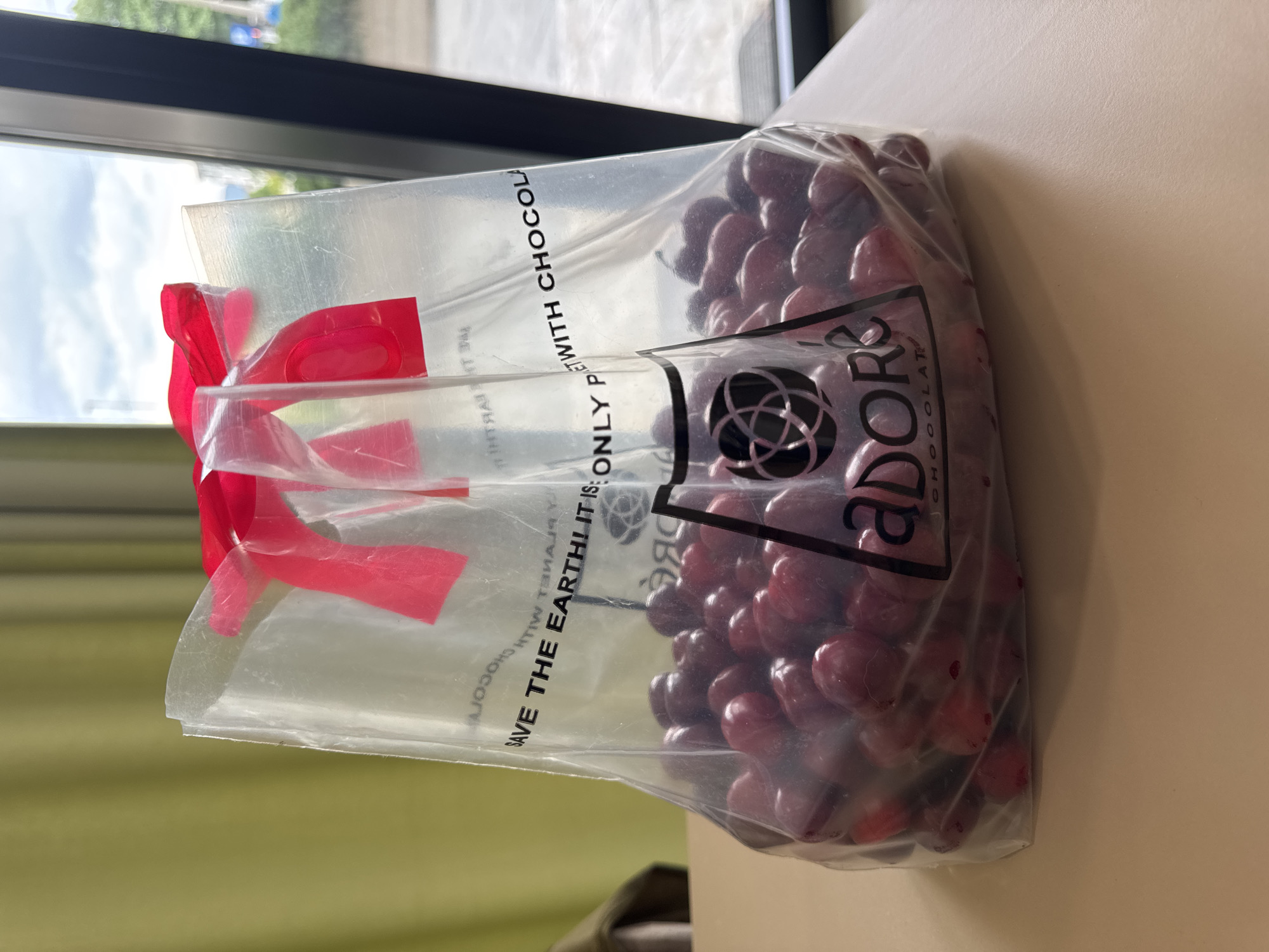 A photograph shows a bag of Adore chocolate on a beige surface, likely a table. The bag is transparent plastic with a red ribbon closure. The branding "adore CHOCOLATE" is visible, along with a circular design. The bag is filled with what appear to be chocolate-covered cherries. The text "SAVE THE EARTH! IT IS ONLY P ET WITH CHOCOLA" is printed on the bag. A window is visible in the background, with a view of trees and buildings, suggesting an indoor setting. The image does not provide any identifying information for Podgorica, Montenegro.