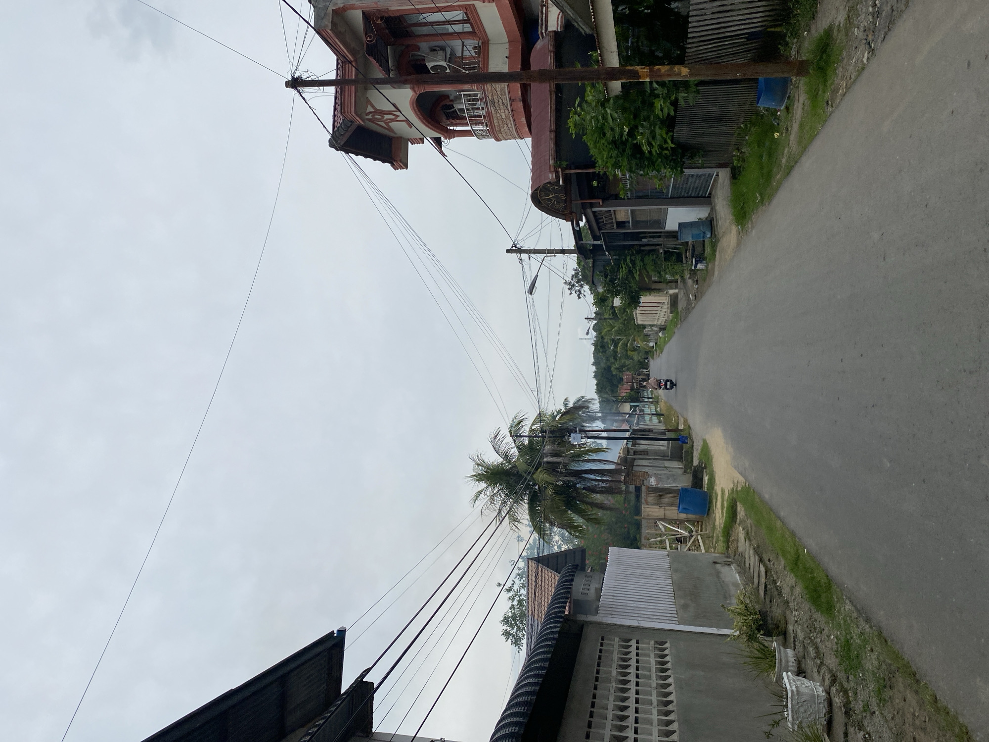 The image depicts a residential street in Lhokseumawe, Indonesia, under an overcast sky. The perspective is from the middle of the paved road, looking straight ahead. On the left side of the street, a gray wall with a decorative pattern of rectangular openings forms the boundary of a property. Several potted plants are visible in front of this wall. Further down the left side, a house with a dark, corrugated roof and a tiled roof is partially visible. Overhead, a dense network of power lines stretches across the sky, supported by utility poles.
On the right side of the street, a multi-story house with cream-colored walls and red trim stands out. It features an arched balcony with decorative metal railings and an air conditioning unit mounted on its facade. Adjacent to this house, other residential structures with varying roof styles and fencing are present. Lush green trees and foliage are interspersed throughout the scene, contributing to a tropical feel.
Further down the road, a lone individual is riding a motorcycle, heading away from the camera. The road itself is asphalt, showing signs of wear. Two blue barrels, likely for water storage or waste, are placed on the side of the road, one on the left and another on the right. The background shows more houses and vegetation, with a gentle slope indicating the terrain. The overall impression is that of a quiet, somewhat undeveloped suburban or semi-rural area within Lhokseumawe.
