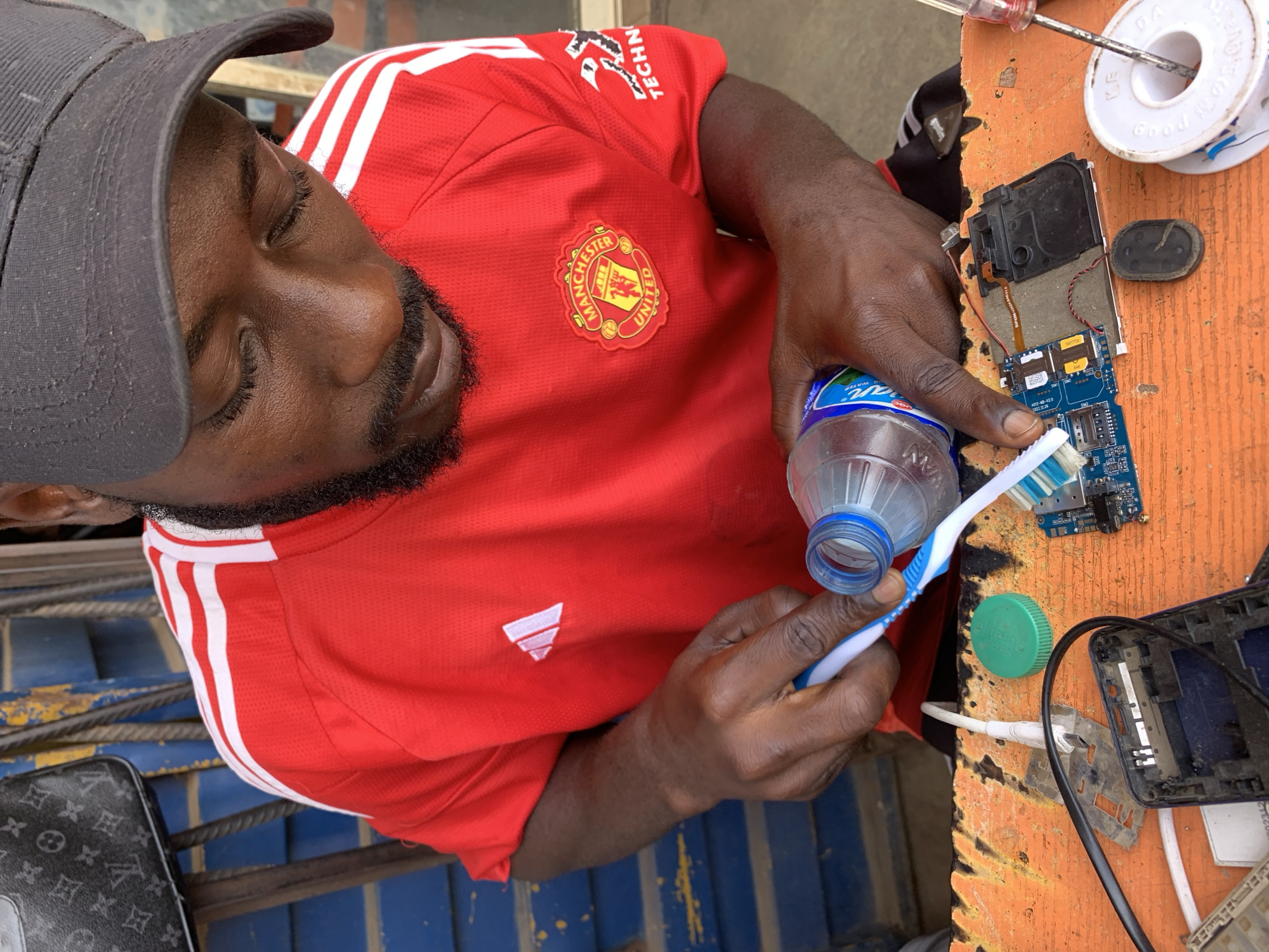 A man wearing a red Manchester United t-shirt and a dark gray baseball cap is using a toothbrush and a water bottle to clean the circuit board of a cell phone. The scene is taking place on a cluttered orange workbench. In the background, a blue metal structure is visible, suggesting the location might be outdoors or in a simple workshop. The location is Rafin Par, Nigeria.
