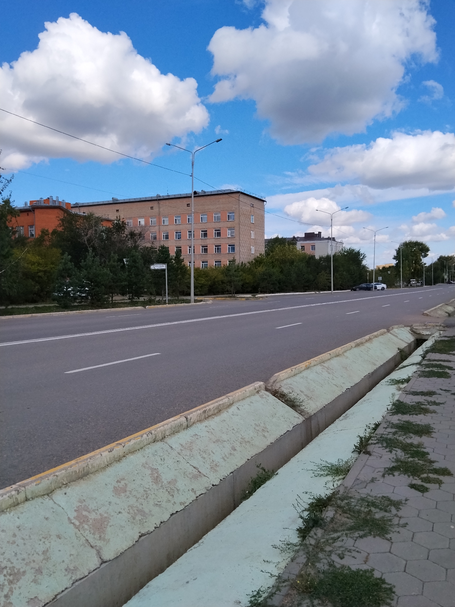 The image depicts a wide asphalt road in Kokshetau, Kazakhstan, with multiple lanes and clear white lane markings. On the left side of the road, there are several multi-story brick buildings, some appearing to be residential or institutional, interspersed with green trees. A street lamp is visible on this side.
On the right side of the road, a concrete drainage ditch runs parallel to the sidewalk. The ditch has a worn, light green painted interior and is partially overgrown with weeds. The sidewalk is paved with hexagonal gray tiles. Beyond the ditch and sidewalk, more trees and the sky are visible. The sky is a bright blue with large, fluffy white cumulus clouds. In the distance, more buildings and street lamps line the road, suggesting an urban or suburban environment. A few cars are visible further down the road, including a white and a dark-colored vehicle. A signpost with text in Cyrillic script is also present near the trees on the left. The overall scene suggests a typical day in an urban setting under a cloudy sky.