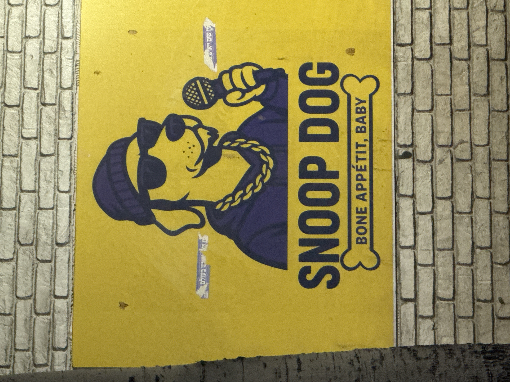 The image is of a poster for "Snoop Dog Bone Appetit, Baby" which features a cartoon dog wearing sunglasses and a beanie holding a microphone and wearing a gold chain. The poster is yellow with blue and black elements. The poster is adhered to a brick wall. There are Hebrew stickers on the poster. The photograph was taken outdoors in Tel Aviv, Israel.