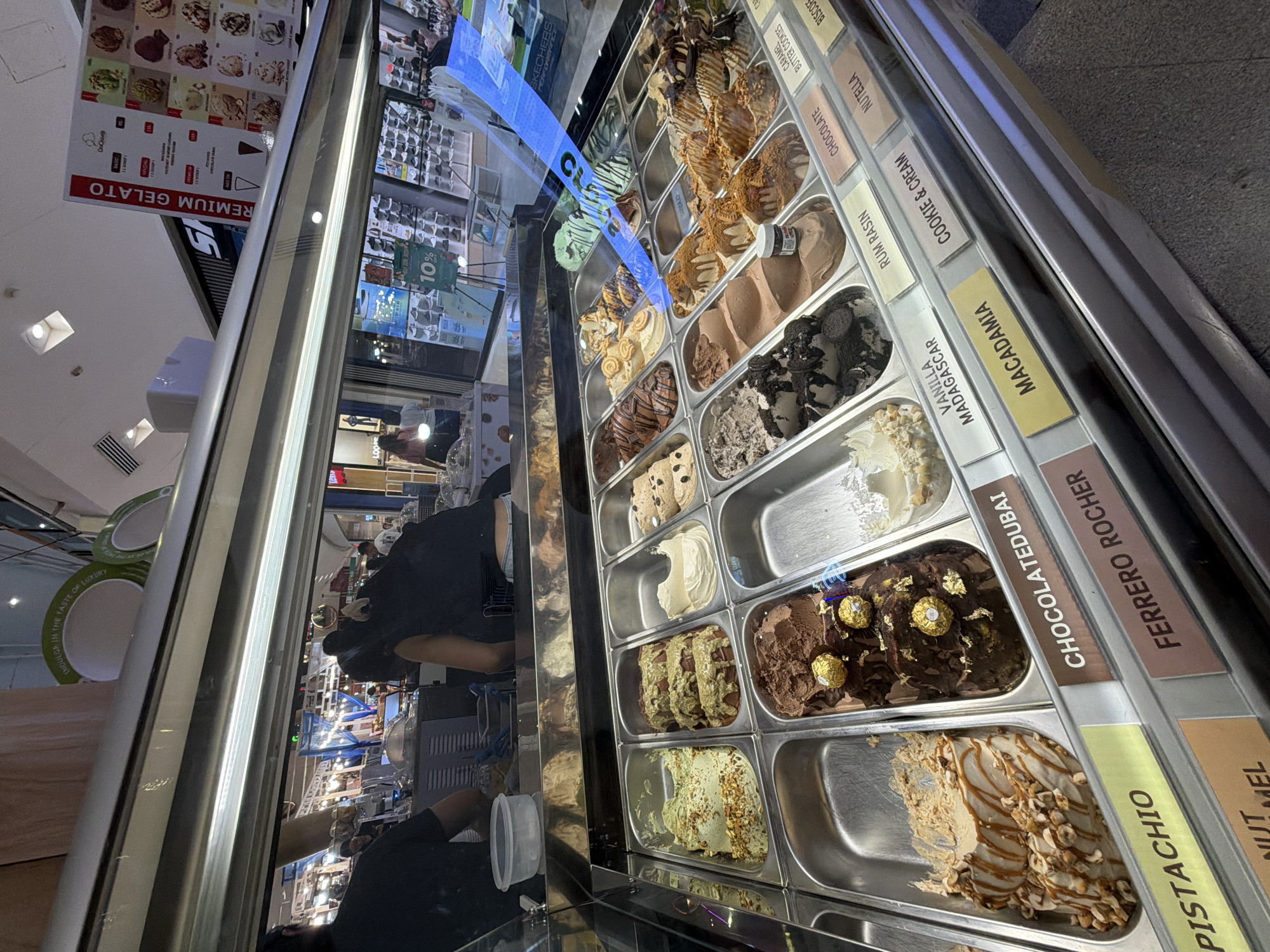A gelato display case is featured in the foreground of the image, containing numerous flavors of gelato. The gelato is presented in rectangular stainless steel containers, with labels indicating the flavors. Some of the visible flavors include Pistachio, Chocolatedubai, Ferrero Rocher, Vanilla Madagascar, Rum Rasin, Cookie & Cream, Macadamia, Chocolate, Nutella, and Biscuit. Behind the gelato case, two individuals are visible, appearing to be staff members. One person, wearing a black t-shirt, is standing behind the counter, and the other, also in black, is seen from the back. In the background, various shops and displays are visible, suggesting an indoor shopping environment. A menu board is mounted on a slanted surface above the gelato case, listing gelato flavors and prices, with the text "PREMIUM GELATO" prominently displayed. The overall scene appears to be an ice cream parlor located within a mall or commercial area.