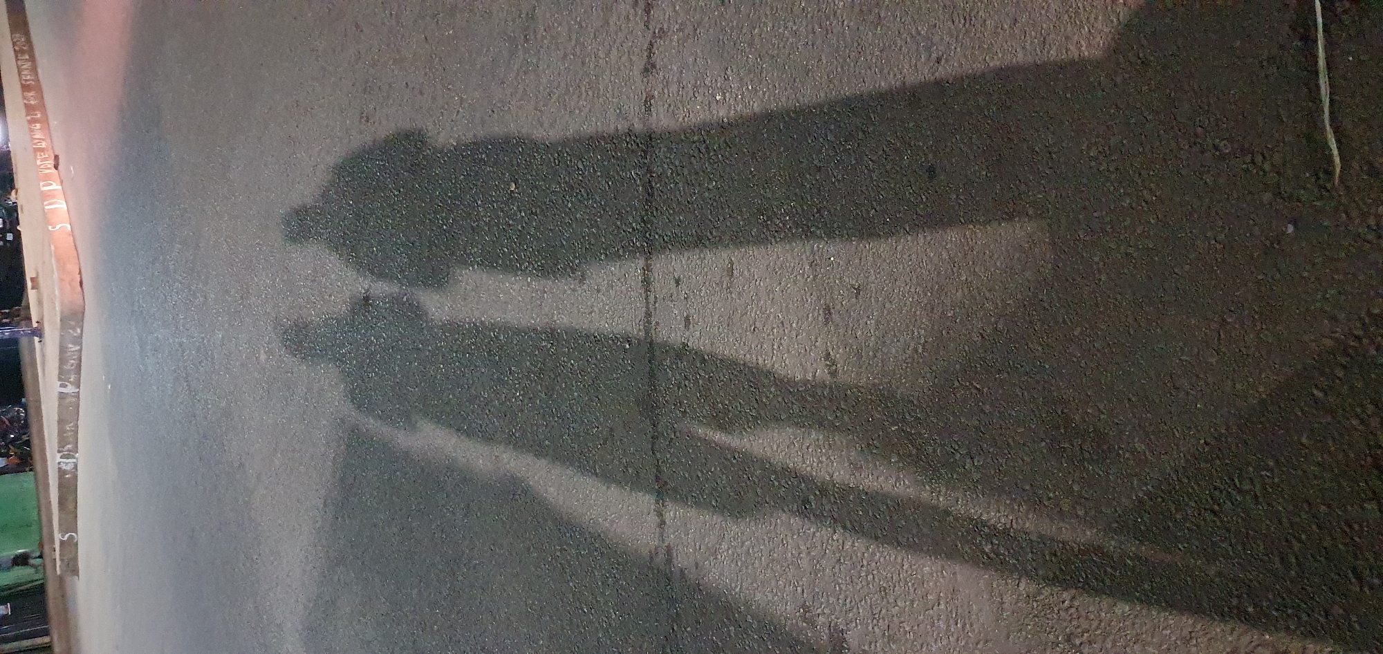 In Barakin Angundi, Nigeria, a low-angle, full-shot photograph captures the elongated shadows of two individuals projected onto an asphalt road at night. The shadows stretch diagonally from the bottom left to the upper right of the frame, indicating a light source positioned behind and to the left of the subjects. The road surface is dark and textured, with some lighter specks visible, suggesting it is slightly damp or dusty.
In the upper portion of the image, a raised concrete curb runs horizontally, with white lettering faintly visible on its surface. Some of this lettering appears to be "SDP VOTE... FOR SENATE 2023". Behind the curb, indistinct figures and objects are visible, suggesting a street or plaza area with some structures or stalls, possibly a marketplace or gathering spot. The lighting suggests artificial light sources, likely streetlights or vehicle headlights, illuminating the scene from behind the subjects. The overall atmosphere is dimly lit and urban.