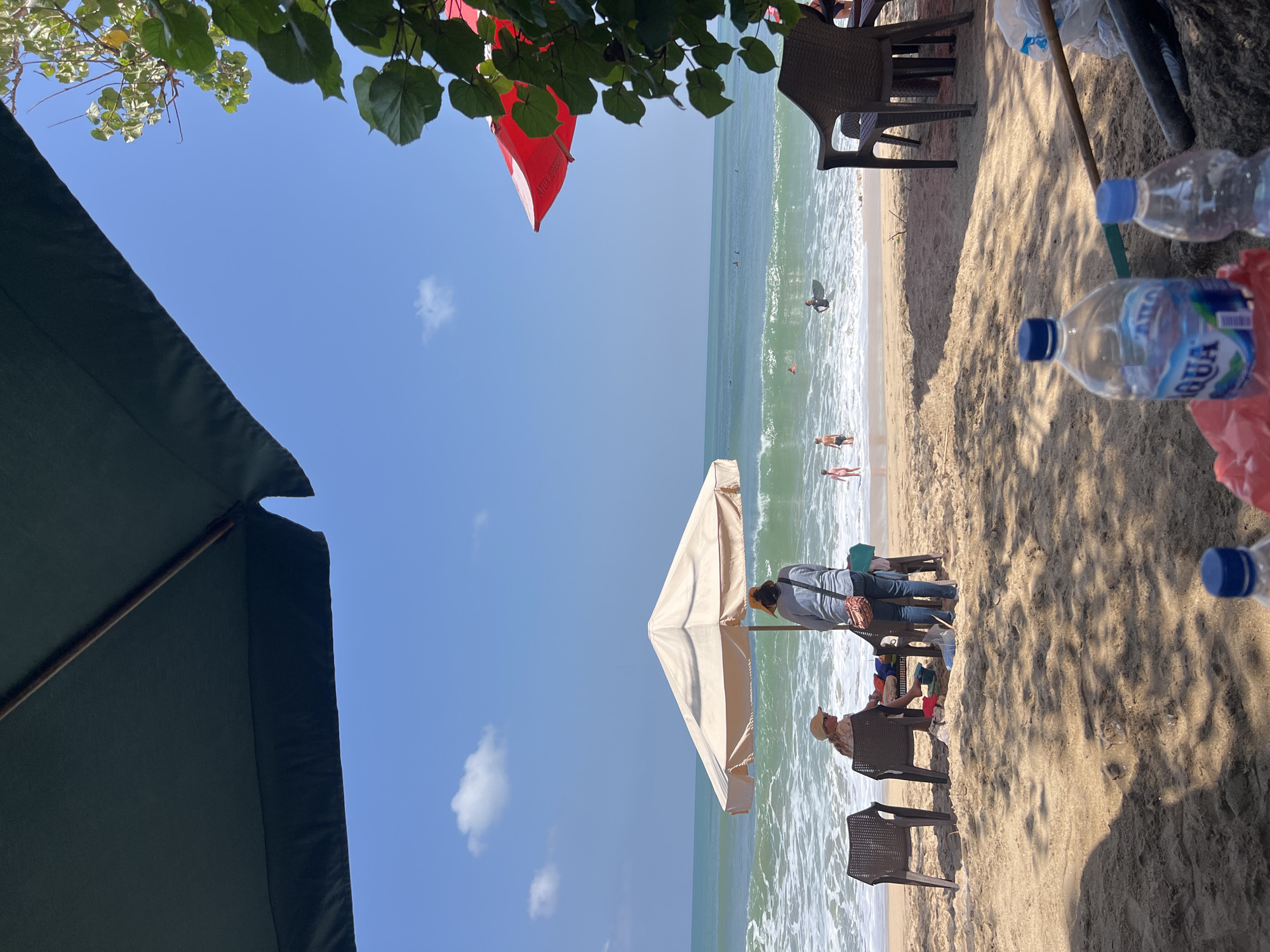 This is a photograph of a beach in Kuta, Indonesia, on a sunny day. The foreground is dominated by sand with dappled shade from an unseen tree. In the middle ground, two beach chairs face the ocean. On the left, a person wearing a hat and sunglasses sits in one chair, appearing to be resting or observing the water. To the right of this person, another individual stands, facing away from the camera, wearing a striped shirt and jeans, with a bag slung over their shoulder. They are positioned near a table under a light-colored umbrella.
In the background, the ocean is visible with moderate waves breaking on the shore. Several people are in the water; some are wading, while others appear to be surfing or bodyboarding. The water is a vibrant green-blue. Further out, more surfers can be seen in the distance. The sky is clear blue with a few scattered white clouds. The presence of umbrellas, chairs, and people in swimwear indicates this is a popular tourist area. The overall scene is one of a relaxed beach day, with recreational activities taking place in the water.