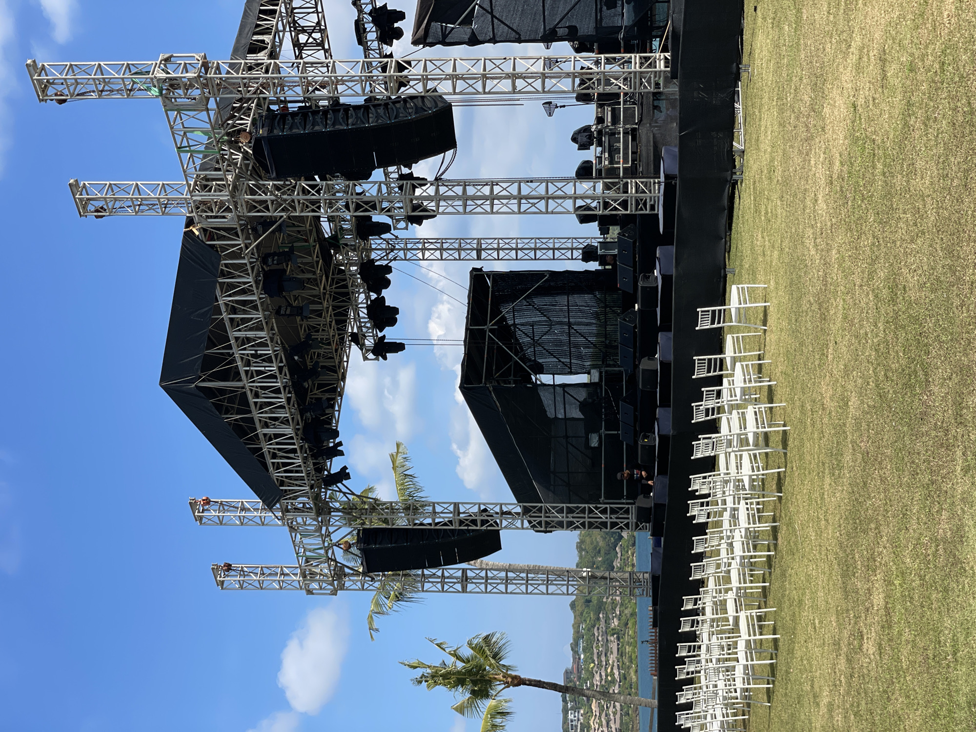 This image captures an elaborate outdoor stage setup, likely for a concert, performance, or large event, situated on a grassy area beside a body of water.
**Subjects:**
The primary subject is a large, complex stage structure built from interlocking metal trusses. This framework supports numerous large black speakers, typically line arrays, and various lighting fixtures. Parts of the stage, including backdrops and equipment covers, are draped in black fabric or screens. On the green grass in front of the stage, multiple rows of white chivari-style chairs are neatly arranged, indicating an audience area. Several tall palm trees are visible, with their fronds extending into the scene, some appearing near or integrated with the stage structure. In the background, a calm blue body of water, possibly an ocean or bay, is visible, with a distant landmass featuring green hills and what appear to be buildings or resort structures along the coastline. A few individuals, presumably crew members, are sparsely visible around the stage, one particularly clear on the right side of the main stage structure and another higher up on a truss.
**Activity or event taking place:**
The scene suggests either the final stages of setting up for an outdoor event or the aftermath. The stage is fully equipped with sound and lighting, and seating is prepared, indicating readiness for a gathering.
**Notable details:**
The time of day is bright and clear, under a blue sky with scattered white clouds, suggesting daytime or late afternoon. The weather appears sunny and pleasant. The presence of palm trees and the ocean strongly indicate a tropical or coastal location. There is no visible text legible in the image. The stage's design includes two prominent cantilevered truss extensions on either side, likely supporting additional speakers or lighting.