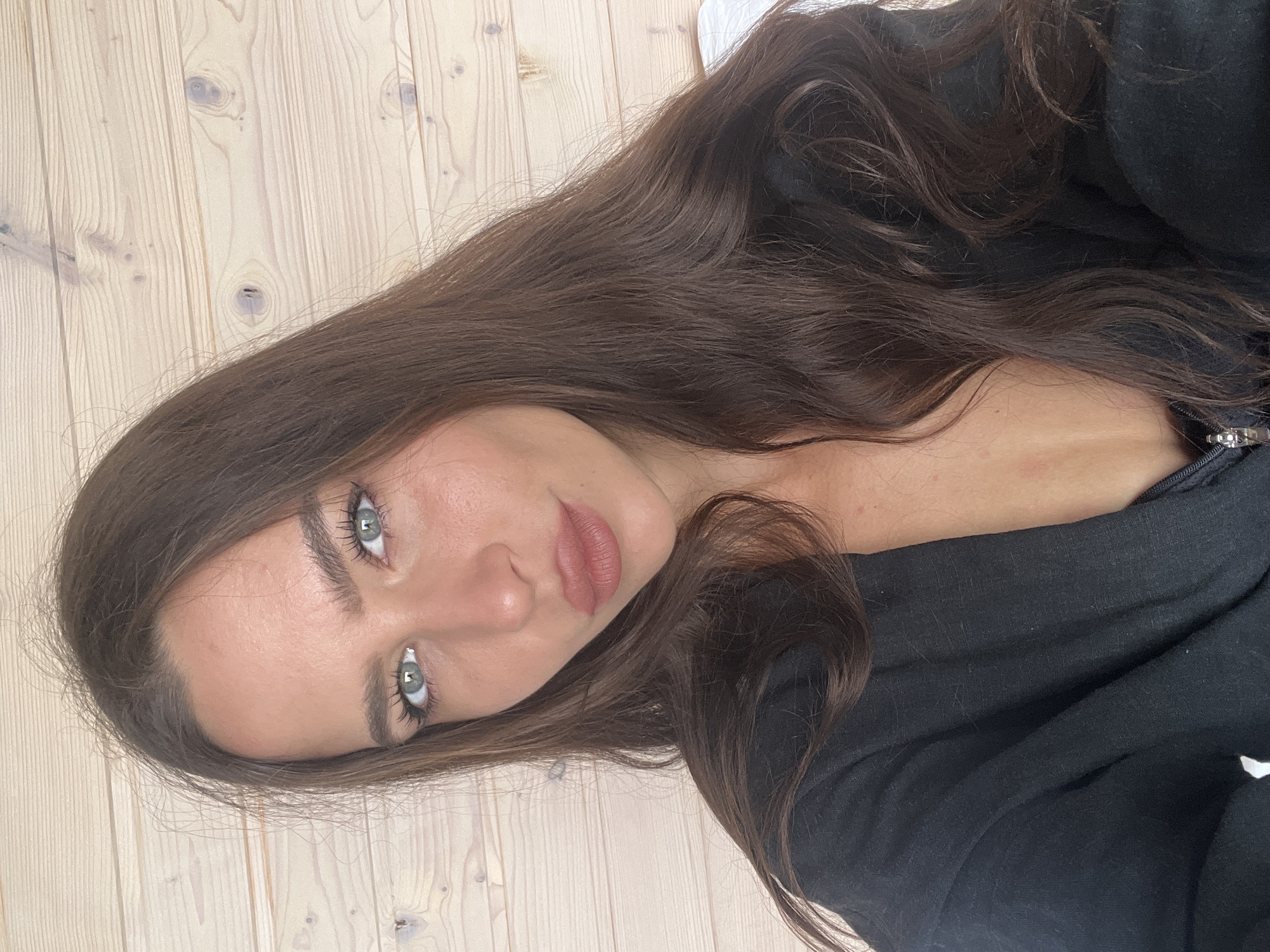 A woman with long, dark brown hair is pictured in a close-up selfie. She has fair skin, light-colored eyes, and her lips are a nude pink. She is wearing a black garment, possibly a shirt or jacket. The background is a light-colored wood surface, likely a floor or wall. The image does not provide any information to identify Zarubino, Russia or any activities, objects or interactions.