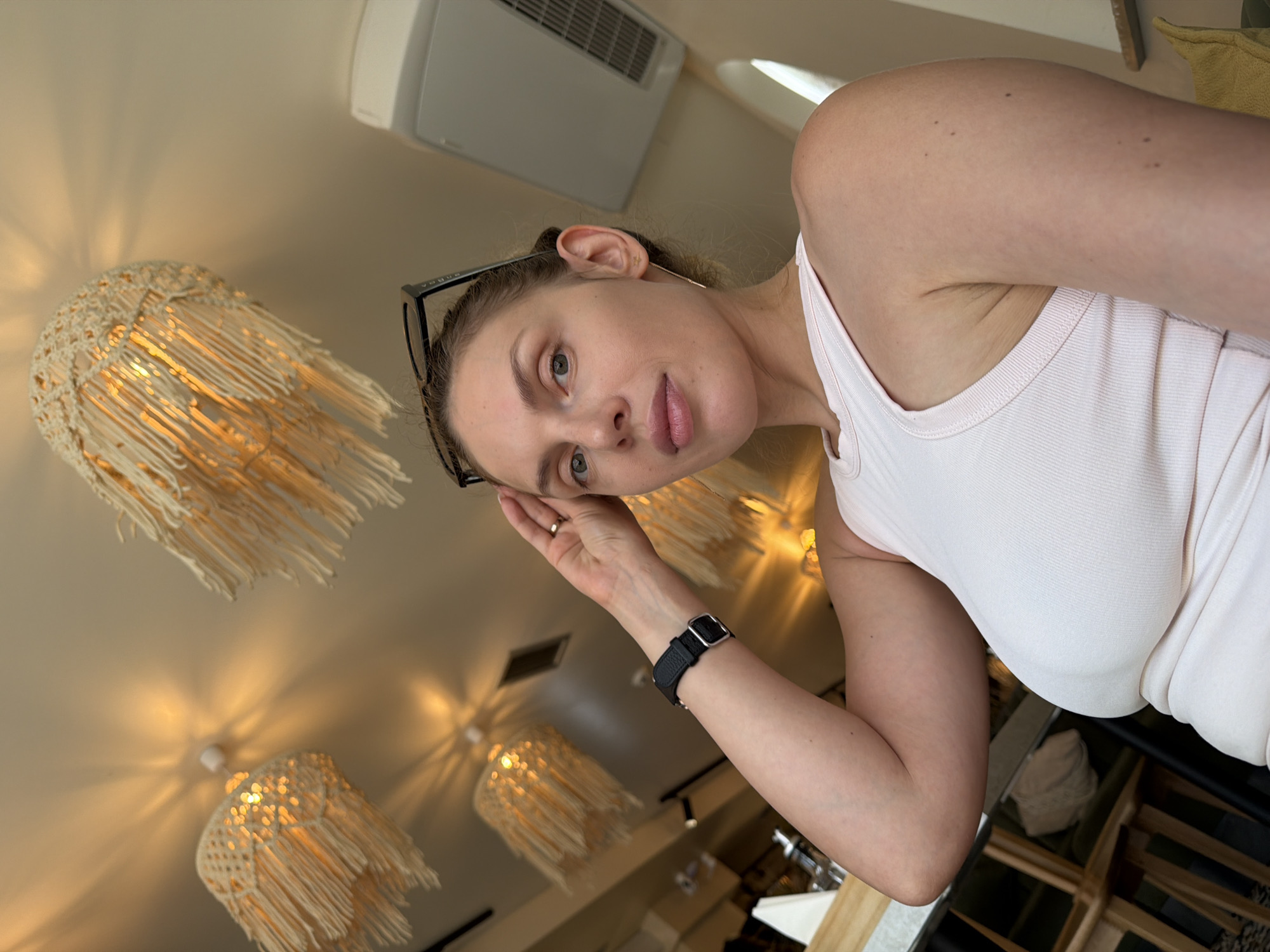 An eye-level, medium shot captures a light-skinned woman with blonde hair tied up and glasses perched on her head. She is wearing a pale pink tank top and looking directly at the camera. Her right hand is raised, touching her forehead, and she has a watch on her left wrist. In the background, several macrame lampshades hang from the ceiling, casting warm light. The scene appears to be indoors, possibly a cafe or restaurant, with hints of furniture and an air conditioning unit visible. The overall lighting is warm and diffused.