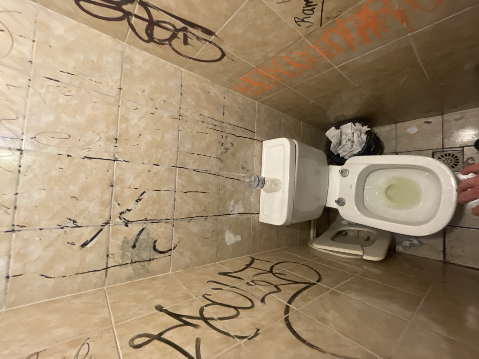 The image shows a tiled bathroom in disarray. Graffiti is scrawled on the walls and floor. A toilet is present, with a water bottle balanced on top of the tank. A wastebasket filled with crumpled paper is next to the toilet. The scene appears to be in a state of neglect or vandalism. The location is Buenos Aires, Argentina.