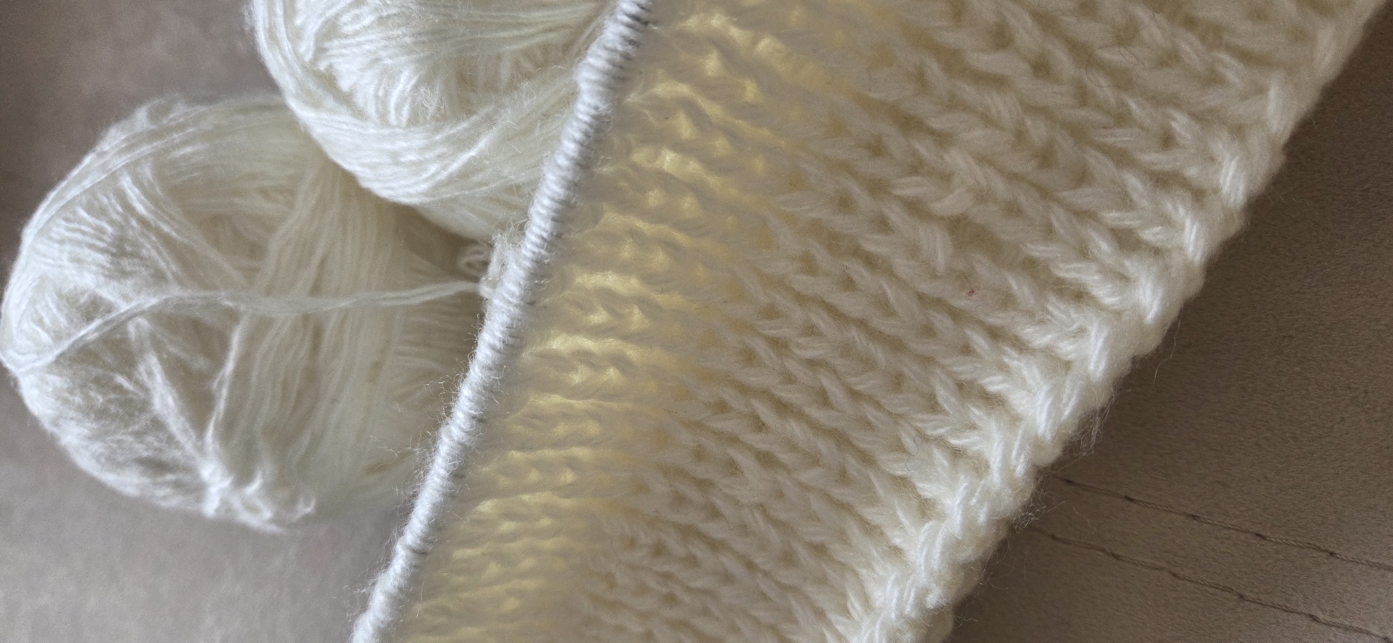 The image displays a close-up view of two skeins of off-white or cream-colored yarn alongside a piece of knitted fabric in the same color.
**Subjects:** The primary subjects are the yarn and the partially completed knitted item. On the left side of the frame, two loose balls or skeins of yarn are visible. The one in the foreground is larger and more clearly defined, showing its loosely wound strands. The knitted fabric, which appears to be in progress, occupies the right side of the image. Its top edge, where stitches would be held on knitting needles (though the needles themselves are not explicitly visible), runs diagonally across the center, separating the yarn from the finished work. The fabric shows a distinct knit stitch pattern, possibly garter stitch or a form of ribbing, characterized by horizontal bars or V-shapes formed by the interlocked loops.
**Activity or event taking place:** The scene suggests the activity of knitting. The presence of the yarn and the partially knitted fabric indicates that a handicraft project is either underway or paused.
**Notable details:** Both the yarn and the knitted fabric are a soft, light, off-white or cream hue. The yarn has a slightly fluffy or fuzzy texture, implying it might be wool or a similar soft fiber. The knitted fabric also exhibits a soft texture and its stitches are regular, suggesting careful craftsmanship. The lighting is soft, highlighting the texture of the yarn and the definition of the stitches. The background appears to be a plain, light-brown or beige surface.
**Visible text:** There is no visible text in the image.
