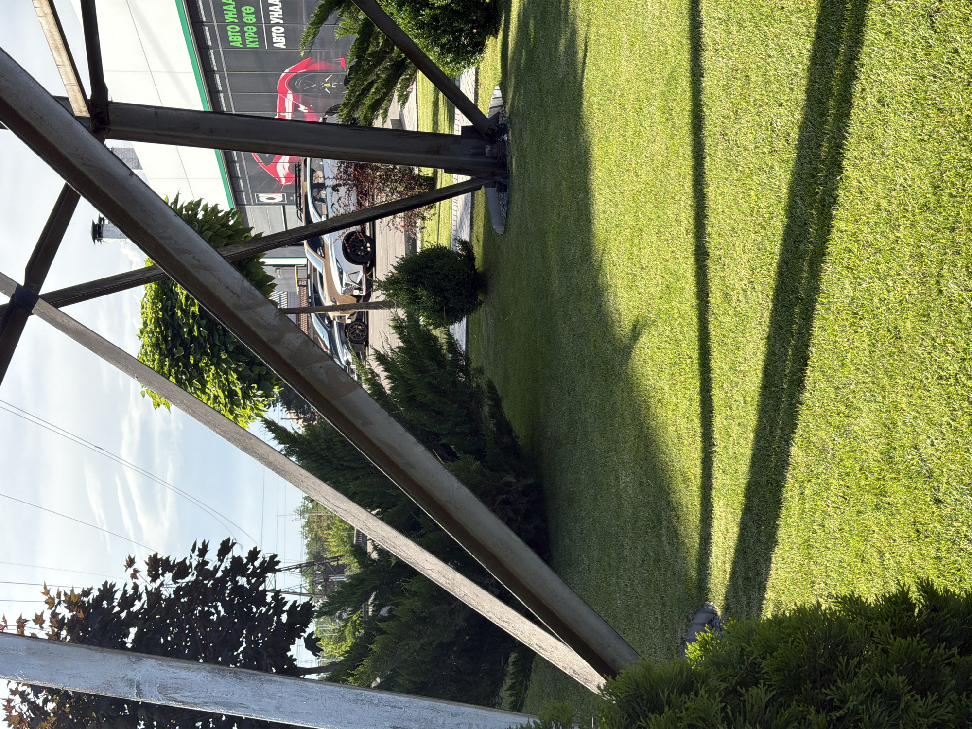 The image shows a close-up of a metal utility pole in Bishkek City, Kyrgyzstan. The pole's construction features a lattice of steel beams. Green lawn surrounds the base of the pole, with shadows cast across the grass. Beyond the pole, a building with a sign in what appears to be a Cyrillic language is visible, along with a row of parked cars. Small bushes and trees are scattered around the base of the pole. The sky is visible through the metal structure.