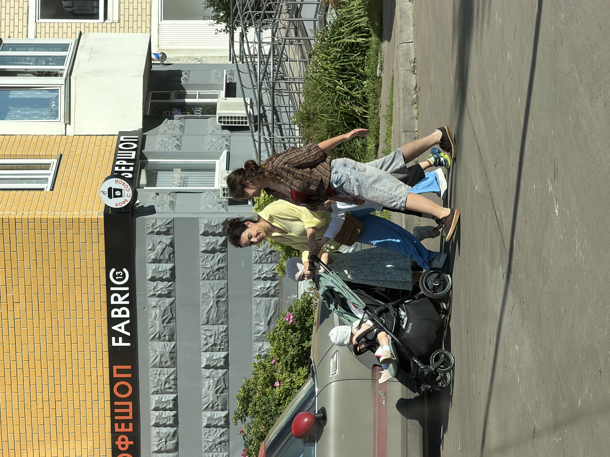 In Moscow, Russia, on a sunny day, two women and two children are walking on a sidewalk next to a street. The woman on the left wears a yellow blouse and blue pants, pushing a stroller with a baby. The woman on the right, in a plaid shirt and denim shorts, walks beside her. The stroller contains a baby in a white hat and a toddler in a blue and white dress. A car is parked on the left side of the street. Behind them, a building with a yellow brick facade and a gray stone lower section has signs that read "ОФЕШОП FABRIC" and a coffee cup logo with "КОФЕ С СОБОЙ" and "ВЕРШОП". There is also a ramp with metal railings to the right of the building. Lush green bushes with pink flowers are visible behind the car. The ground is paved with asphalt.