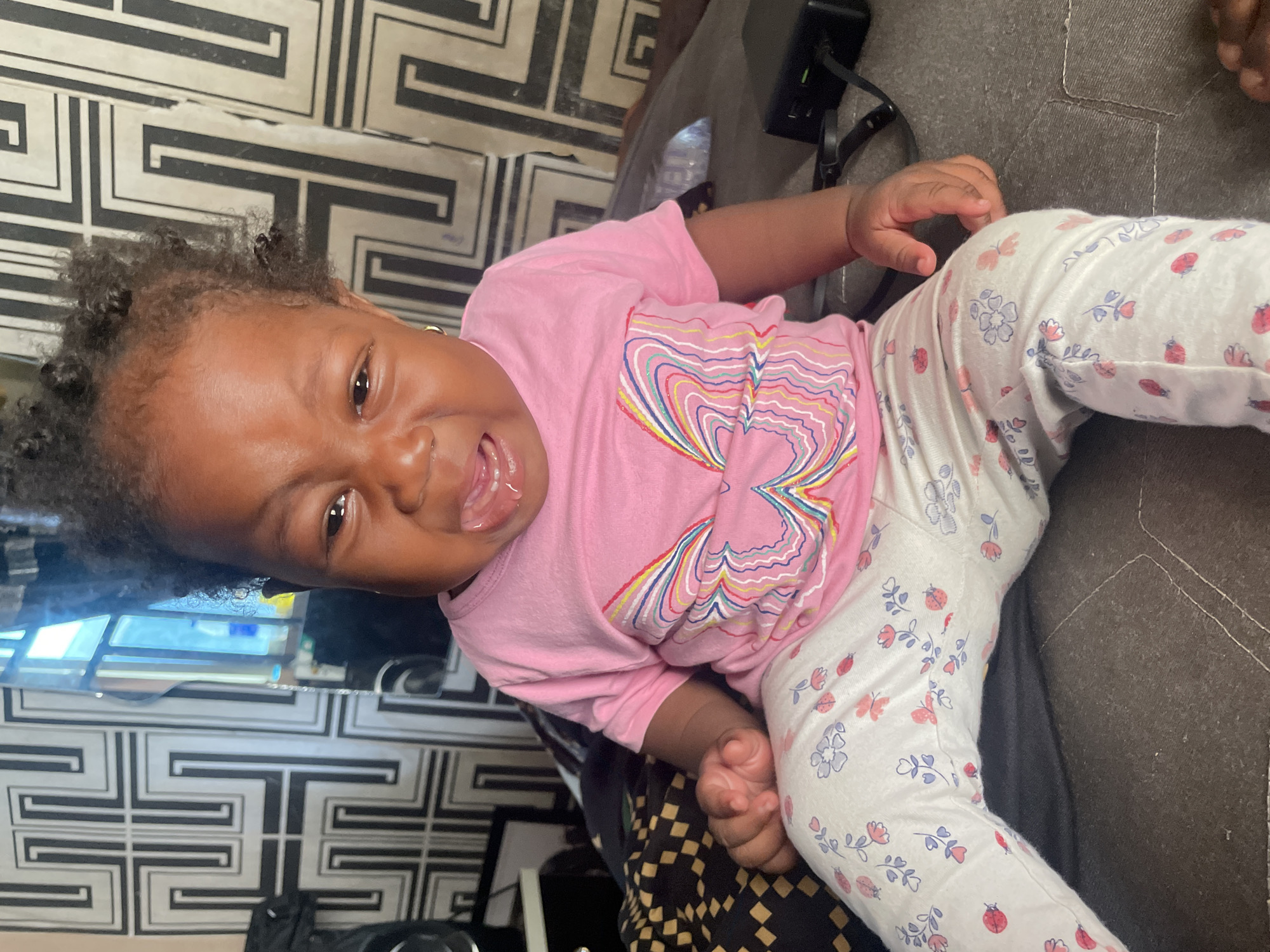 A baby is lying on a dark-colored couch. The baby is wearing a pink long-sleeved shirt with a colorful heart design and patterned white leggings with floral and ladybug motifs. The baby is facing the camera with a wide open mouth and slightly squinted eyes. Behind the baby, there is a wall with a black and white geometric pattern. There is also a dark-colored object with wires attached, possibly a power strip.
The location of the image is unknown and cannot be determined as Lokoja, Nigeria.