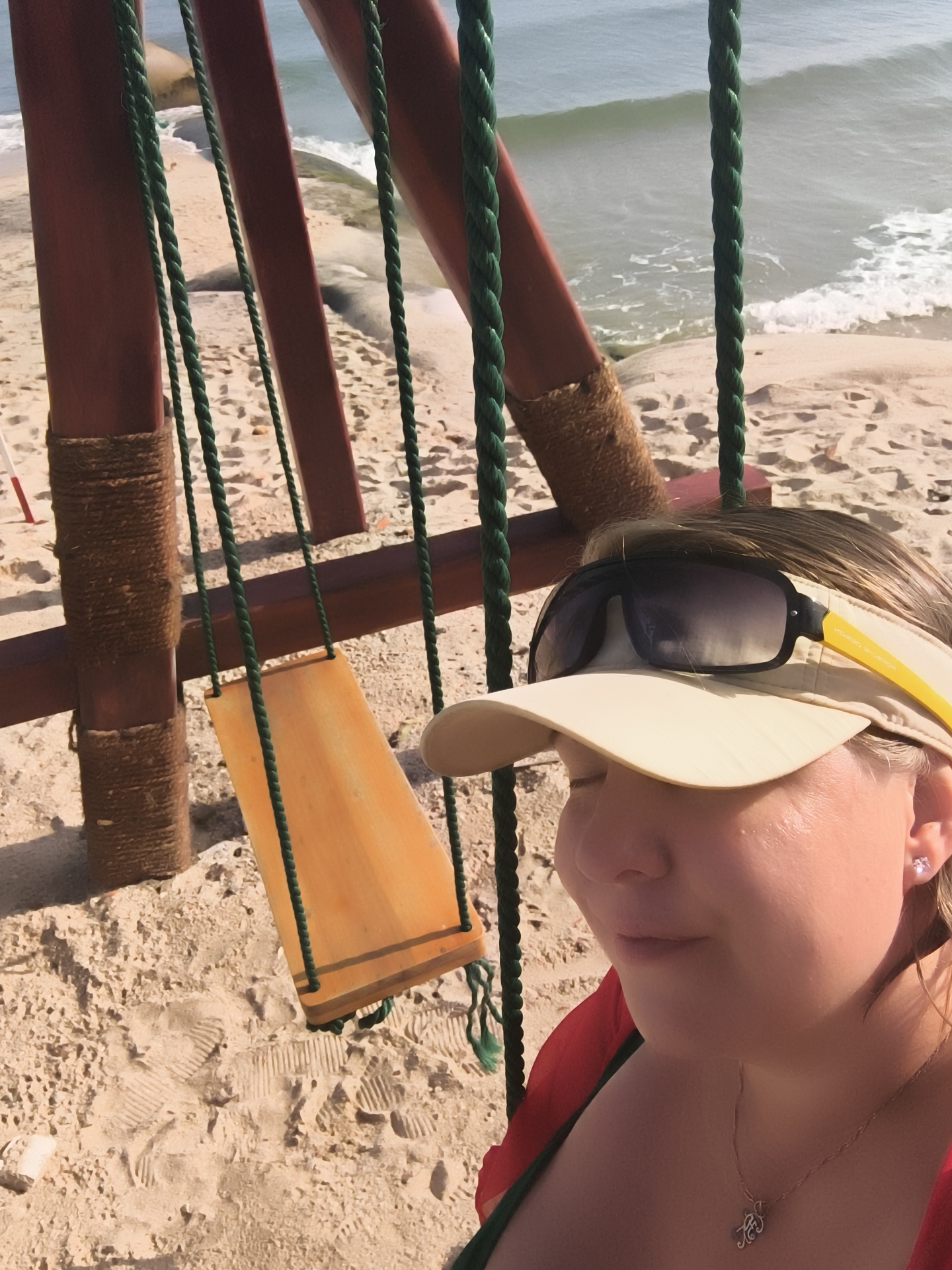 A woman wearing a visor and sunglasses is seen in a close-up shot from the shoulders up, on a sandy beach. Behind her, a wooden swing with green ropes is partially visible. The ocean and waves can be seen in the background. The scene appears to be in Phu Thuy Ward, Vietnam.