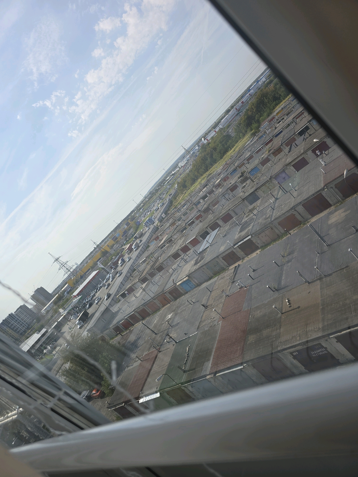 The image, taken from an elevated perspective, shows a vast expanse of connected garages with their doors closed. The garages appear to be constructed from concrete and metal, with some doors painted in various colors like red, blue, and brown. Numerous streetlights are visible above the garages, casting shadows on the ground. In the background, a road with parked cars and a few trucks can be seen, alongside some trees and buildings. A yellow industrial-looking structure and power lines are also present. The sky is partly cloudy with a light blue hue. The scene is likely a garage complex in an urban or semi-urban area. Based on visual cues and common urban planning in Russia, this could be a typical garage cooperative. While the specific location in Ulyanovsk, Russia cannot be definitively identified without further information, the image depicts a characteristic urban landscape element found in many Russian cities.