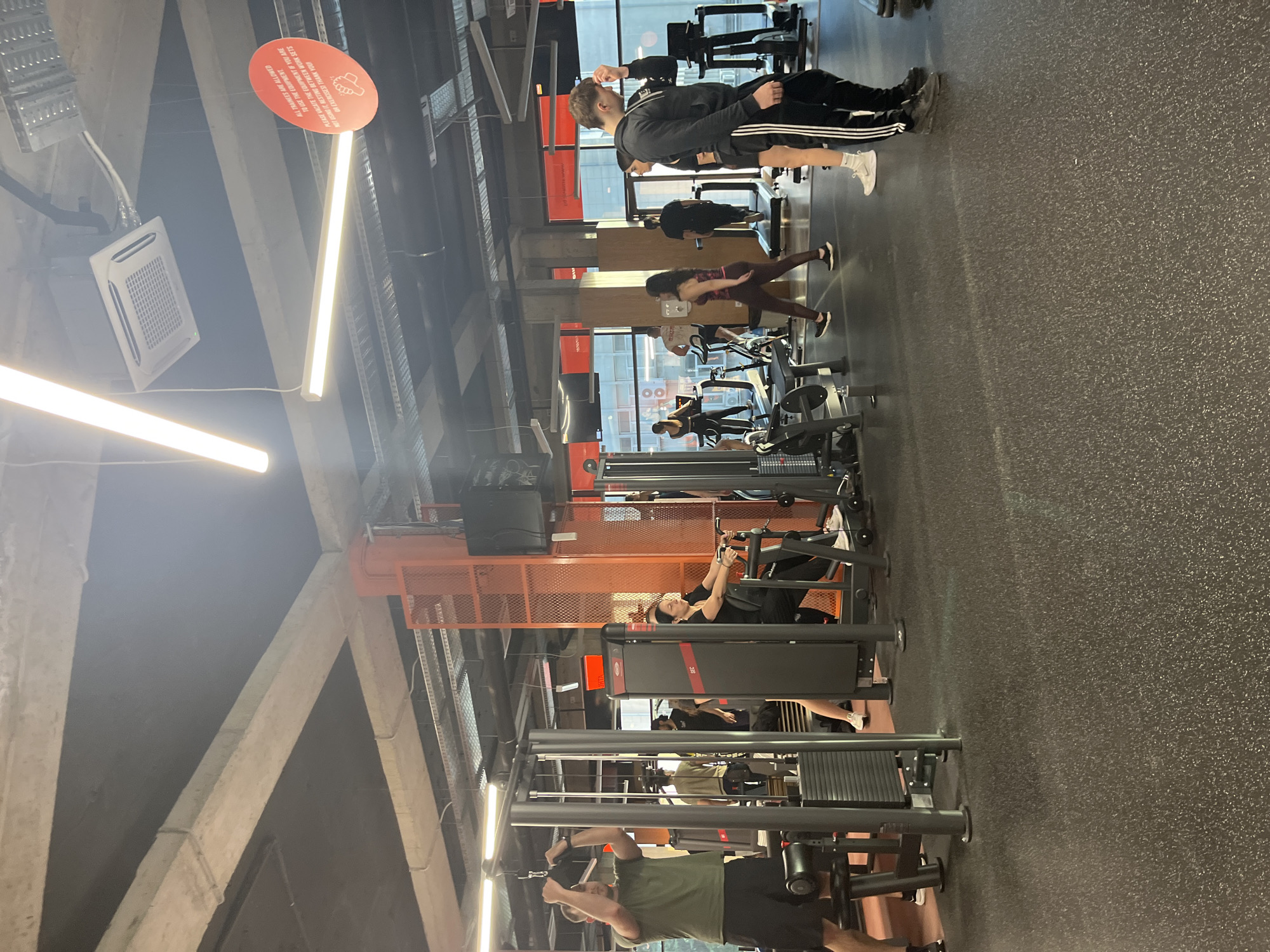 The image shows the interior of a gym in Plovdiv, Bulgaria. Several people are exercising on various machines. In the foreground, a man is using a seated row machine. Further back, a woman is working out on a chest press machine. Other people are using leg press machines and free weights. The gym has an industrial aesthetic with exposed concrete beams and orange-colored metal supports. A sign hangs from the ceiling with the text: "PLEASE VACATE THE EQUIPMENT IF YOU ARE NOT USING IT, RESTING BETWEEN WORK SETS OR EXERCISESST THANK YOU!"