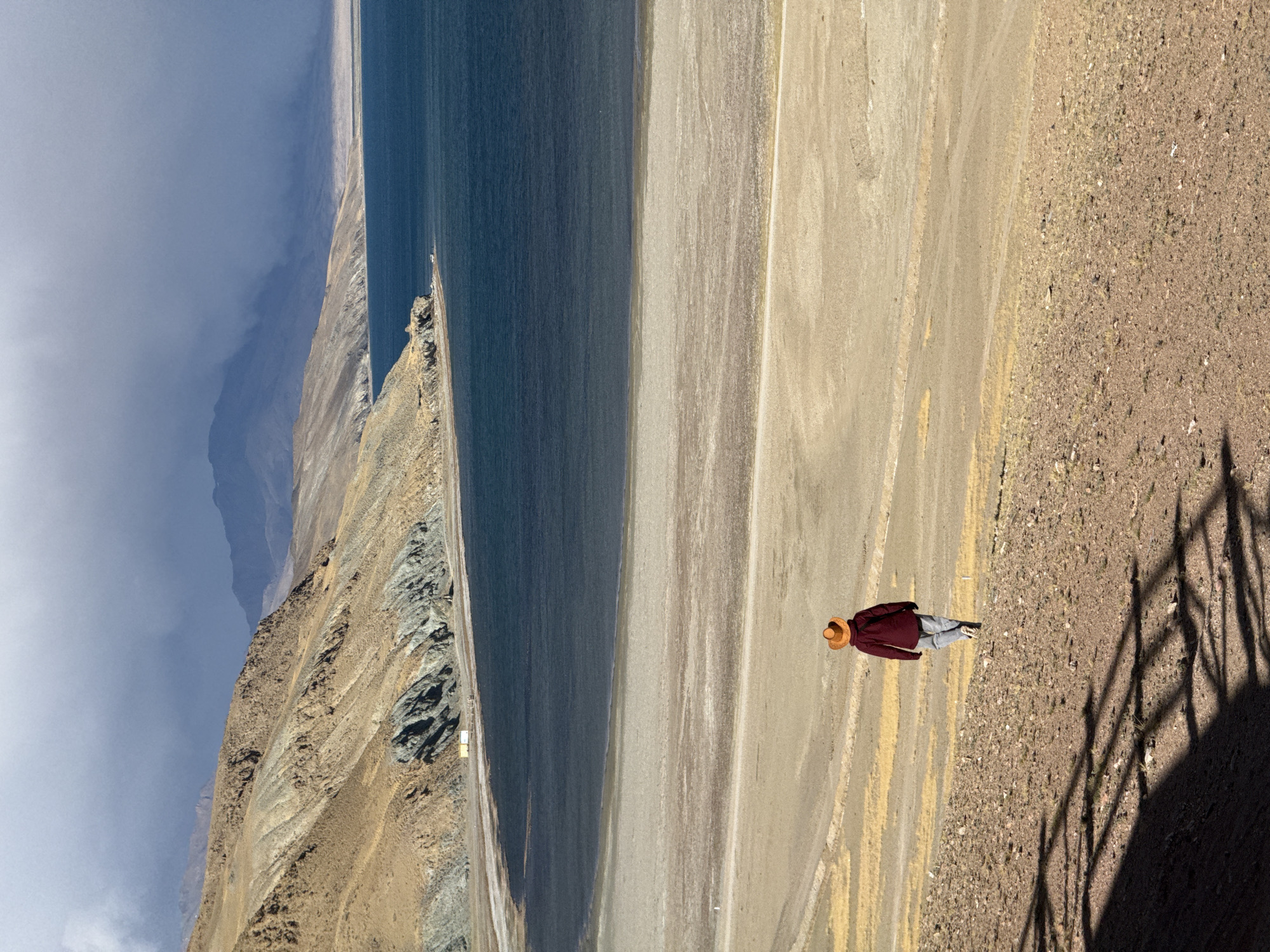 A high-angle, rotated view presents a landscape where a body of water borders a sandy shoreline. Mountains rise in the background, shrouded in clouds. A person wearing a maroon jacket and tan hat stands on the sandy terrain. The shadow of a fence or similar structure is visible on the right side of the frame. The person appears still, looking out at the scenery. No location is listed in this image.