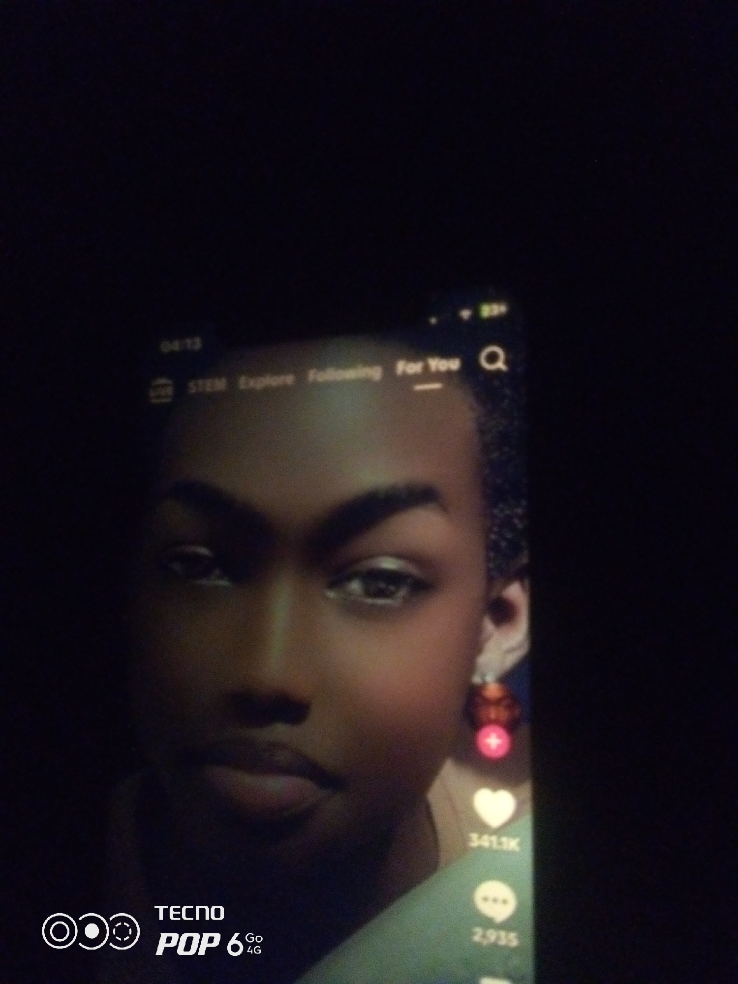 This is a dimly lit, close-up photograph of a person's face, viewed on a mobile phone screen. The person has dark skin and is looking slightly off-camera. Their eyebrows are dark and arched, and they are wearing subtle makeup, including eyeliner and mascara. A small, dangly earring with red accents is visible on their right ear.
The phone screen displays a social media interface, likely TikTok, as indicated by the icons and text visible. At the top of the screen, the time is 04:13, and the battery is at 83%. Navigation options like "STEM," "Explore," "Following," and "For You" are present, along with a search icon. On the right side of the screen, interaction icons are visible: a pink plus sign, a white heart with "341.1K" below it, and a speech bubble icon with "2,935" beneath it.
At the bottom left of the phone screen, a logo for "TECNO POP 6 4G" is displayed. The background of the photograph is predominantly dark, suggesting the surrounding environment is also dimly lit. The overall scene appears to be an indoor setting, with the focus sharply on the phone screen and the person's face. The location is unknown.