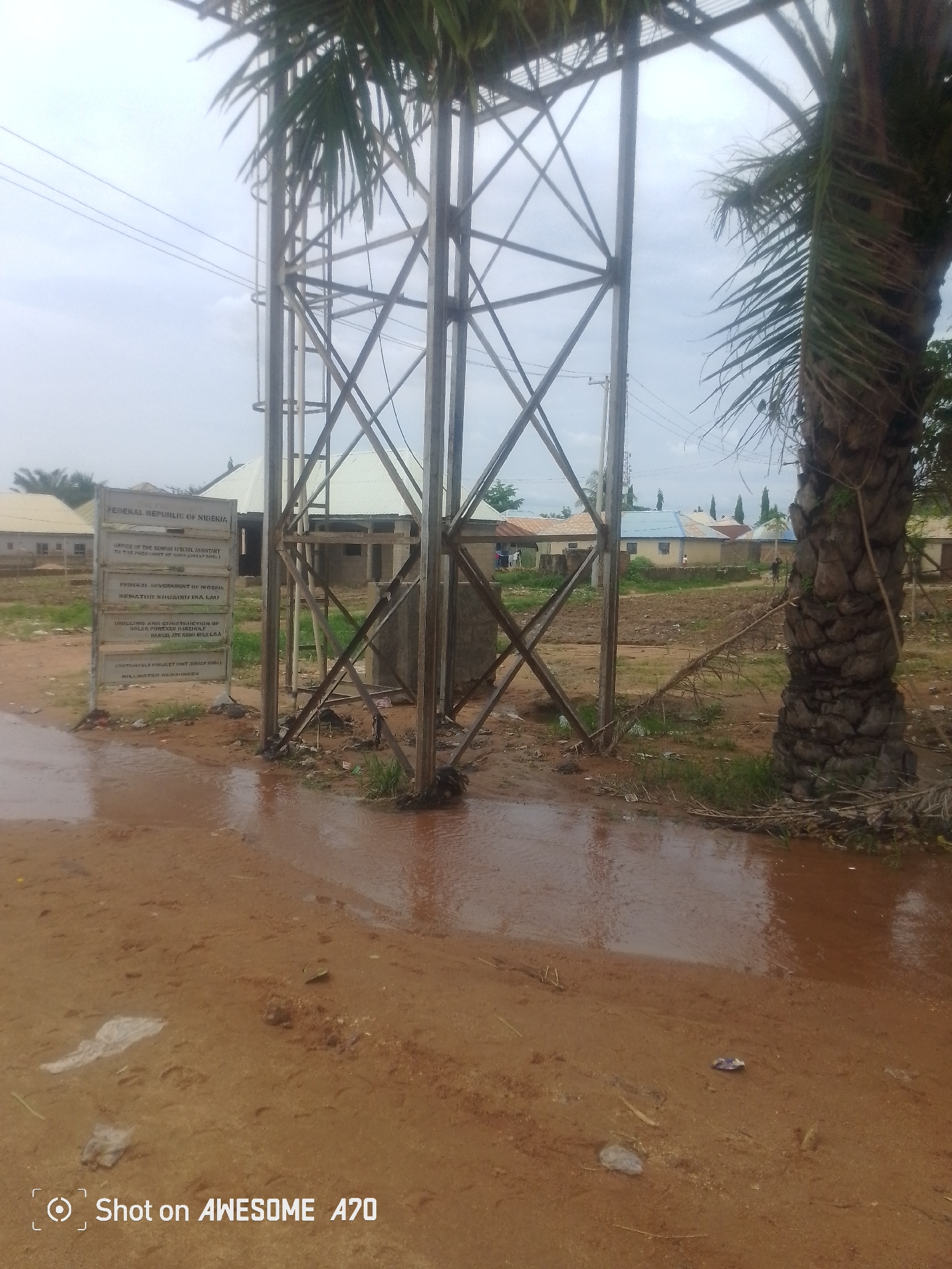 The image depicts an outdoor scene in Jauro Yinu, Nigeria, under an overcast sky. In the foreground, a dirt path is wet and reflects the surroundings, suggesting recent rainfall. To the left, a metal tower structure stands prominently, with palm fronds hanging from a tree to the right. A signpost is located near the base of the tower, displaying text which appears to be government-related. Behind the tower, several buildings with corrugated metal roofs are visible, some appearing to be under construction. Sparse vegetation and debris are scattered across the ground. A few indistinct figures can be seen in the background near the buildings. The overall impression is of an undeveloped or semi-developed area.