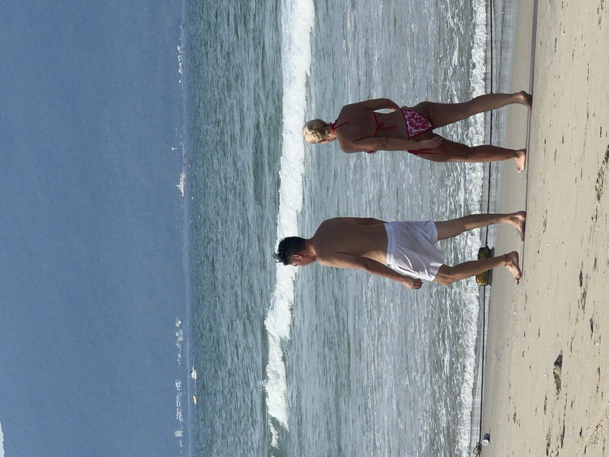 The image depicts two individuals, a woman and a man, at a beach. The woman is wearing a red and white patterned bikini, and the man is wearing white shorts. They appear to be standing or walking on the sandy beach with the ocean in the background. The waves are visible, suggesting it's a day at the seaside.
The scene is set on a beach, with the ocean and waves forming the backdrop for the subjects. The sky appears to be a clear blue, indicating good weather, and the time of day seems to be daytime, likely morning or afternoon given the lighting. The presence of the ocean and the sandy shore points to a coastal location.
The people are casually dressed for a beach environment, and their posture suggests they are engaged in a leisurely activity like walking or standing by the water. There are no specific objects that stand out prominently, apart from the natural elements of the beach and sea. The overall mood appears relaxed and summery.
No visible text is present in the image. The location context of Da Nang, Vietnam, provides further information about the geographical setting of this beach scene.