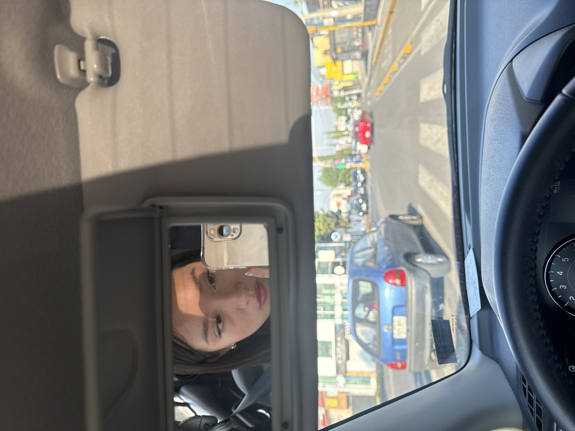 The image is a view from inside a car, presumably the driver's perspective. The car is on a road in Puebla, Mexico, indicated by the license plates of the blue car in front. The road appears to be a city street with several buildings, traffic signals, and other vehicles visible in the background. The driver has a sun visor down, which includes a vanity mirror. Reflected in the vanity mirror is the face of a young woman holding a smartphone, apparently taking a picture. The steering wheel is visible on the right side of the image.
