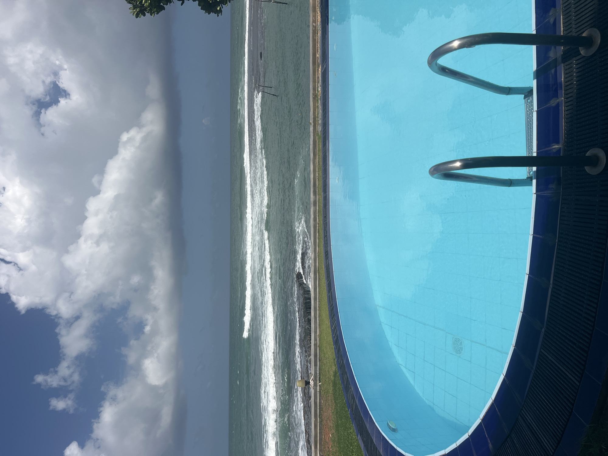The image depicts a scenic view of an outdoor swimming pool with a large body of ocean water in the background. The swimming pool, filled with clear turquoise water, takes up the foreground and is surrounded by a dark blue tiled border and a black grated edge. Two metallic handrails for pool access are visible in the center of the pool. Beyond the pool, a strip of green grass separates it from a rocky shoreline. The ocean is a deep green color with white waves crashing onto the shore and further out into the sea. The sky is a dramatic mix of blue patches and large, dense gray clouds. The location is identified as Goviyapana, Sri Lanka, suggesting a coastal resort or private residence with ocean access. No people are present in the image.