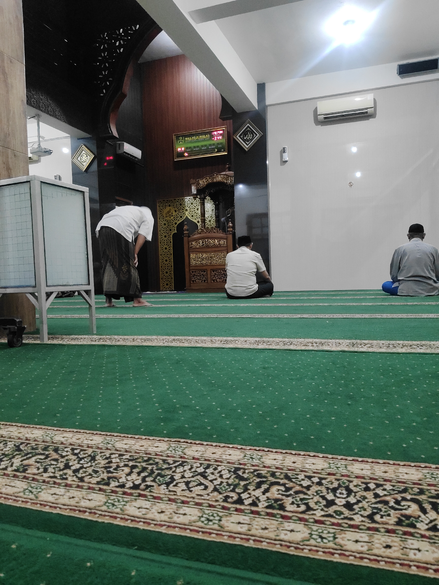 A medium shot captures an interior scene within a mosque. The perspective is low, looking across a green carpet with a decorative border. In the foreground, the patterned border of the carpet is clearly visible. To the left, a whiteboard on a stand is partially visible. Three men are present in the scene. One man, on the far left, is seen from behind, bent over in prayer. He is wearing a white shirt and a dark patterned sarong. In the mid-ground, a second man is seated on the carpet, facing away from the camera, dressed in a white shirt and dark trousers. On the right side of the frame, a third man is also seated on the carpet, facing away from the camera, wearing a light grey shirt and blue trousers. Behind the seated men, a large decorative wooden structure, likely part of the mosque's interior design, is visible. Above this, a digital display shows time and prayer information. The ceiling features dark wood paneling with decorative cutouts. The walls are painted a light color, and an air conditioning unit is mounted on the right wall. The lighting appears to be a mix of overhead lights, with one light source directly above the seated men creating a bright glare.