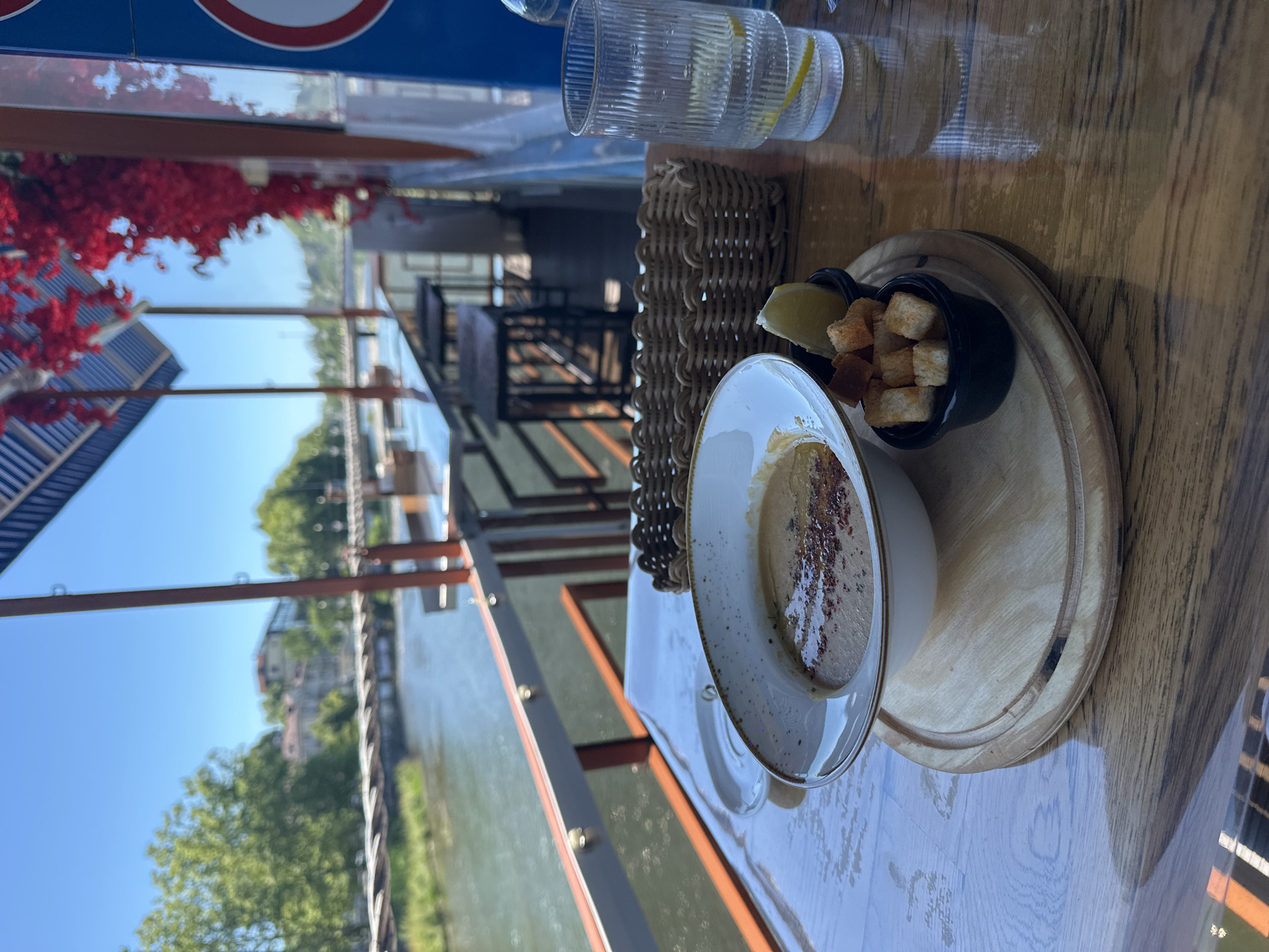 An overhead shot captures a table at a restaurant overlooking a body of water, possibly a river or canal, in Sochi, Russia. The scene is set during daytime under a clear blue sky. On the table, a white bowl filled with a creamy dip, likely hummus, is presented with a sprinkle of red seasoning on top. Beside the bowl, a small black dish holds toasted croutons and a wedge of lemon. The food is arranged on a light wooden serving board. To the left of the food, a woven basket and a ridged glass filled with water and lemon slices add to the table setting. In the background, through large windows, trees and buildings are visible, suggesting an urban or waterside setting. The overall ambiance appears relaxed and leisurely, possibly indicating a meal enjoyed outdoors or in a restaurant with an open-air feel. The light filtering through the windows suggests it is a bright, sunny day.