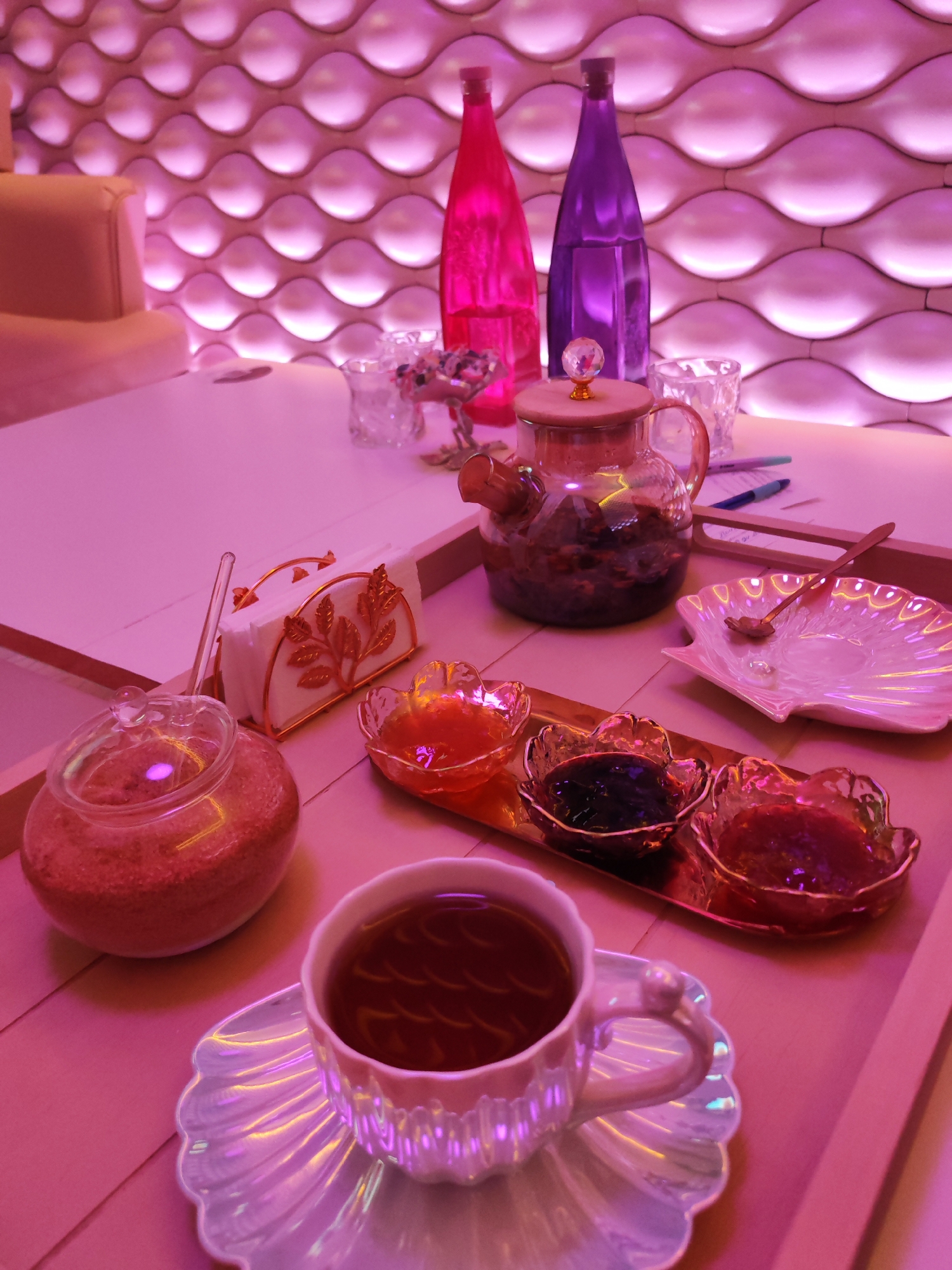 The image depicts a still life of a tea service on a wooden tray. A teacup filled with tea is positioned in the foreground. Behind it is a sugar jar filled with sugar. A metal stand of napkins. A row of three small dishes, each containing a different color jam. Further back, a clear glass teapot filled with tea. Behind the teapot, two decorative colored bottles, one pink and one purple. Two small glass candles are visible. The background is a wall covered in wave-like geometric panels illuminated with pink light. The location is possibly Izhevsk, Russia.