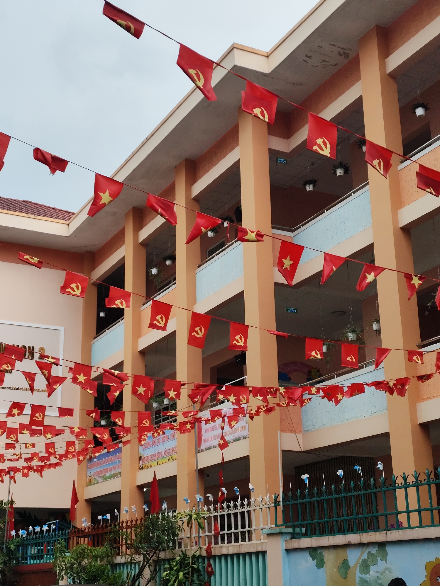 The image depicts a building in Da Lat, Vietnam, likely a school or government building, adorned with numerous red flags. Some flags bear a yellow star (Vietnam's national flag), while others feature a yellow hammer and sickle, symbolizing communism. The building has multiple stories with peach-colored columns and light blue balconies. Hanging plants are visible on the balconies. In the foreground, a fence separates the building from the street, and there are decorative windmills and some greenery. A banner with Vietnamese writing is visible, indicating a festive occasion or commemoration. The sky is overcast.
