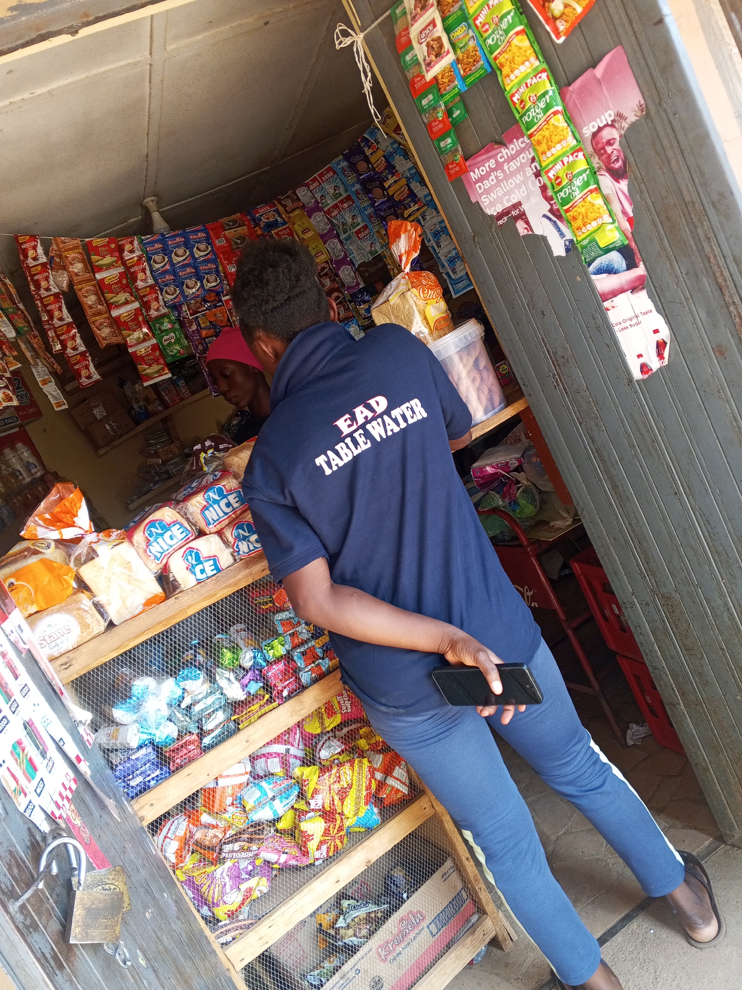A medium-shot, slightly angled photograph captures a person in blue athletic wear standing inside a small shop in Jos, Nigeria. The individual wears a dark blue t-shirt with the words "EAD TABLE WATER" printed on the back. They hold a smartphone in their left hand.
Inside the shop, a woman with a red head covering stands behind a display shelf stocked with bread, snacks, and other goods. The shelves are made of wire mesh. Bags of chips and other small items hang from the ceiling and walls.
Outside the shop, part of a metal door is visible. The door has advertisements and signs posted on it, including branding for "MINI PACK Power Oil" and "Cola Original Taste". The floor is a light-colored stone. A red plastic chair is partially visible behind the person in the blue shirt.