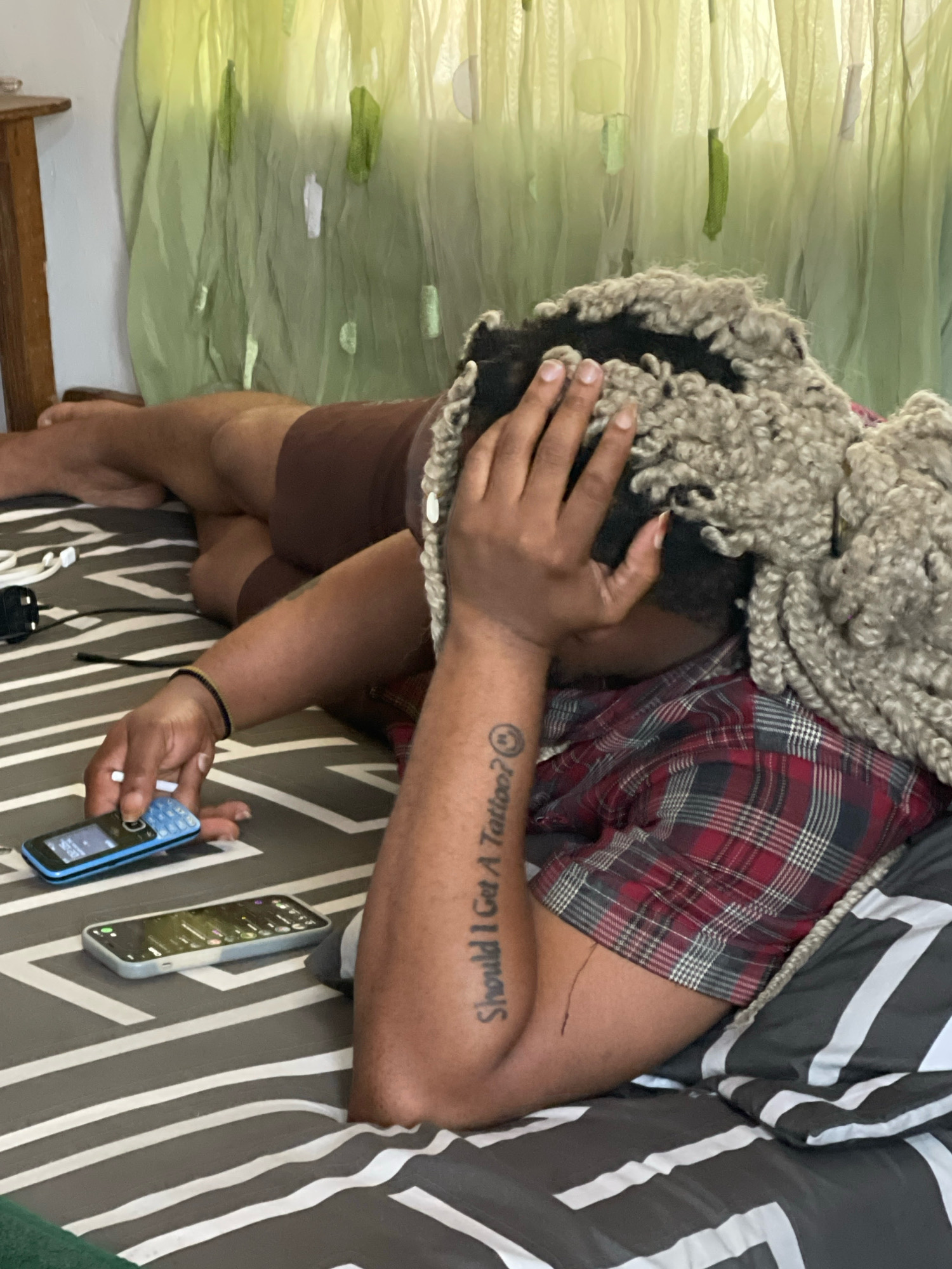 A person with voluminous grey, curly hair lies on a bed covered with grey and white striped bedding. They are wearing a red and black plaid shirt and brown shorts. Their left arm is bent, with their hand resting on their forehead, obscuring their face. A tattoo on their right forearm reads "Should I Get A Tattoo?" followed by a smiley face emoji. Two mobile phones are on the bed: a blue feature phone to the left, and a modern smartphone to the right. White earbuds and a black power adapter are also visible near the phones. The background features a light green, sheer curtain. This scene is taking place in a bedroom in Jos, Nigeria.
