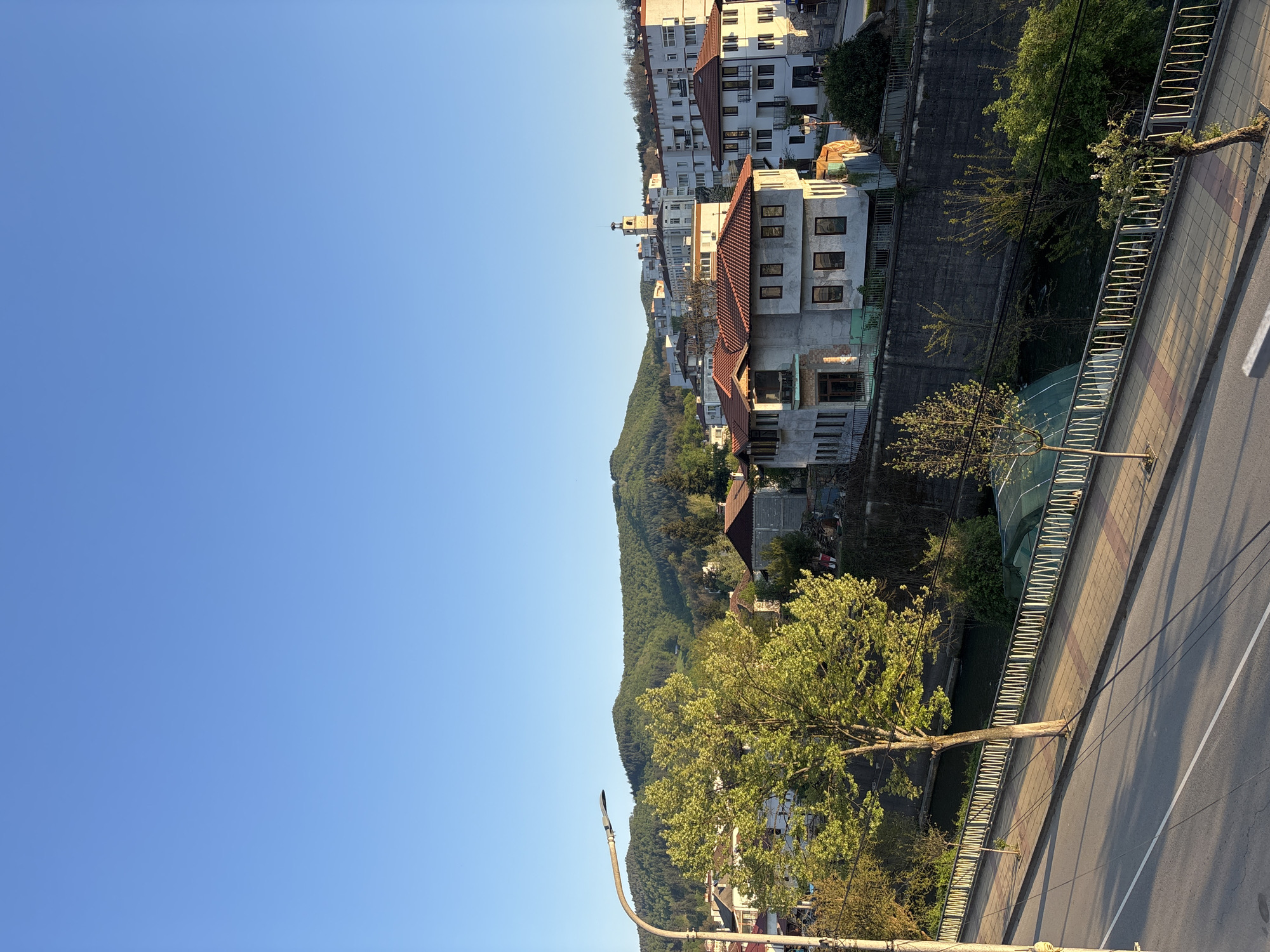 The image depicts a view of Zlatograd, Bulgaria, looking toward a hillside of trees and buildings under a clear blue sky. A street or road runs along the bottom right of the frame, bordered by a metal railing. Trees grow along the hillside, and multiple buildings are visible. The style of architecture is typical of urban areas in Bulgaria.