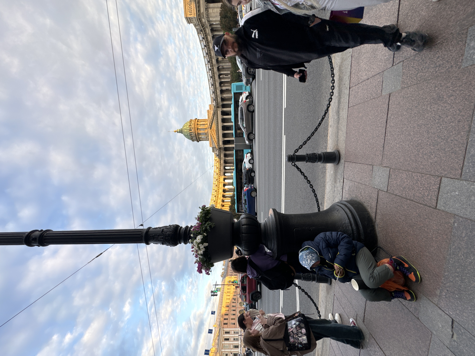 The image depicts a street scene in Saint Petersburg, Russia, likely in front of the Kazan Cathedral, evidenced by the domed building in the background. In the foreground, a large, ornate black lamppost with a planter of flowers stands prominently. To the left of the lamppost, three young women are gathered, one of whom is holding a beverage. Adjacent to the lamppost and further to the left, a person in a blue puffer jacket and a patterned headscarf is seated on an orange bag, looking at a phone and holding a white cup. To the right of the lamppost, a man with a beard, wearing a black Puma tracksuit and a baseball cap, is walking and holding a phone. Behind the people and the lamppost, a street with traffic, including a blue bus, cars, and a red SUV, is visible. The sky is partly cloudy. The pavement is made of reddish-brown granite. A metal chain fence runs along the sidewalk, separating it from the street.