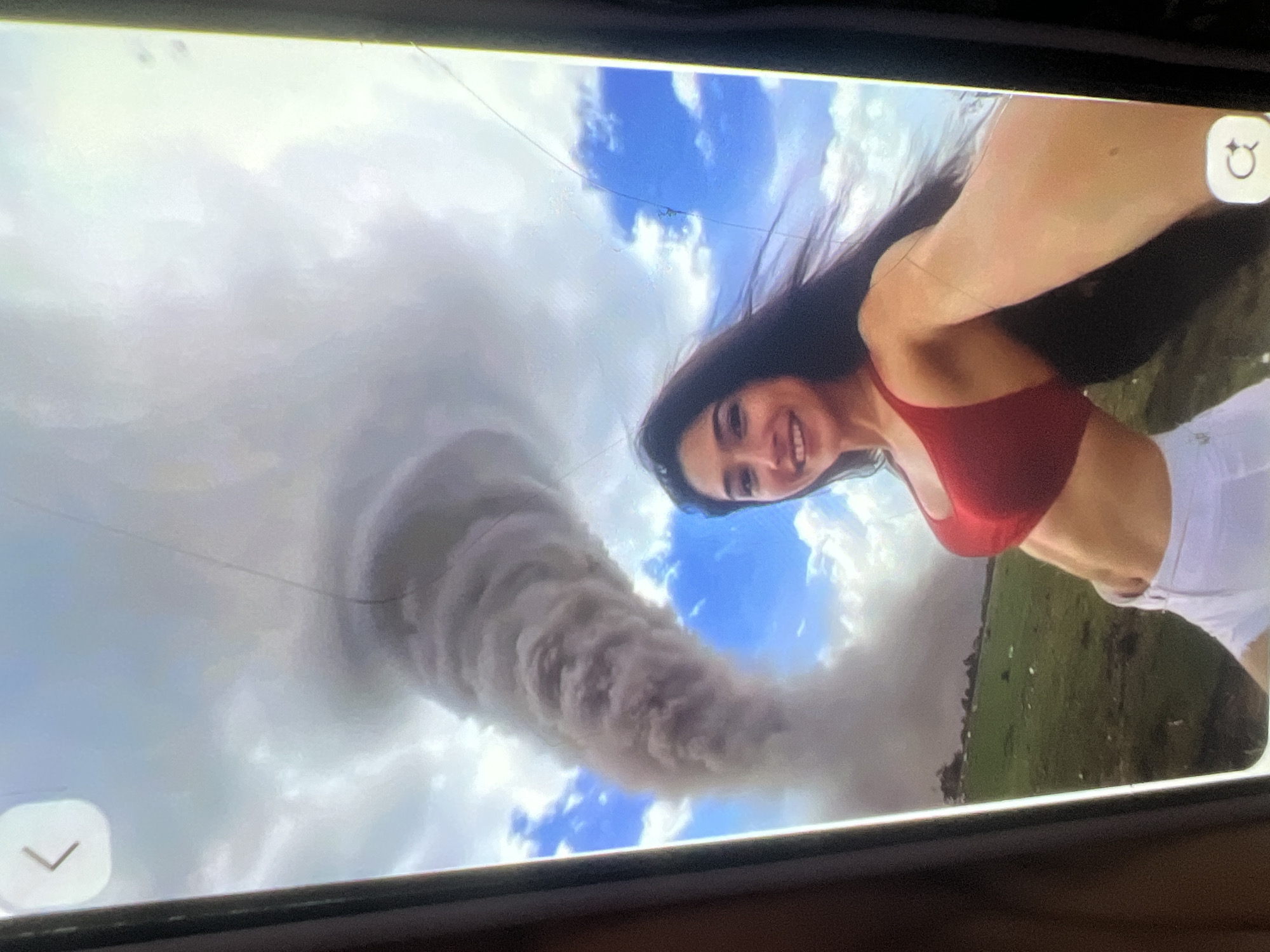 The image depicts a selfie taken by a woman in a red bikini top and white shorts. Behind her, a large dust devil or small tornado is visible in a field under a cloudy blue sky. The woman is smiling and looking at the camera. The scene appears to be in a rural area with green fields and some scattered debris. The overall impression is of a dramatic natural event occurring in the background while the subject poses for a photo. The location specified, Kara-Dıykan, Kyrgyzstan, is not explicitly visible in the image, but the landscape could be consistent with a rural area in that region.