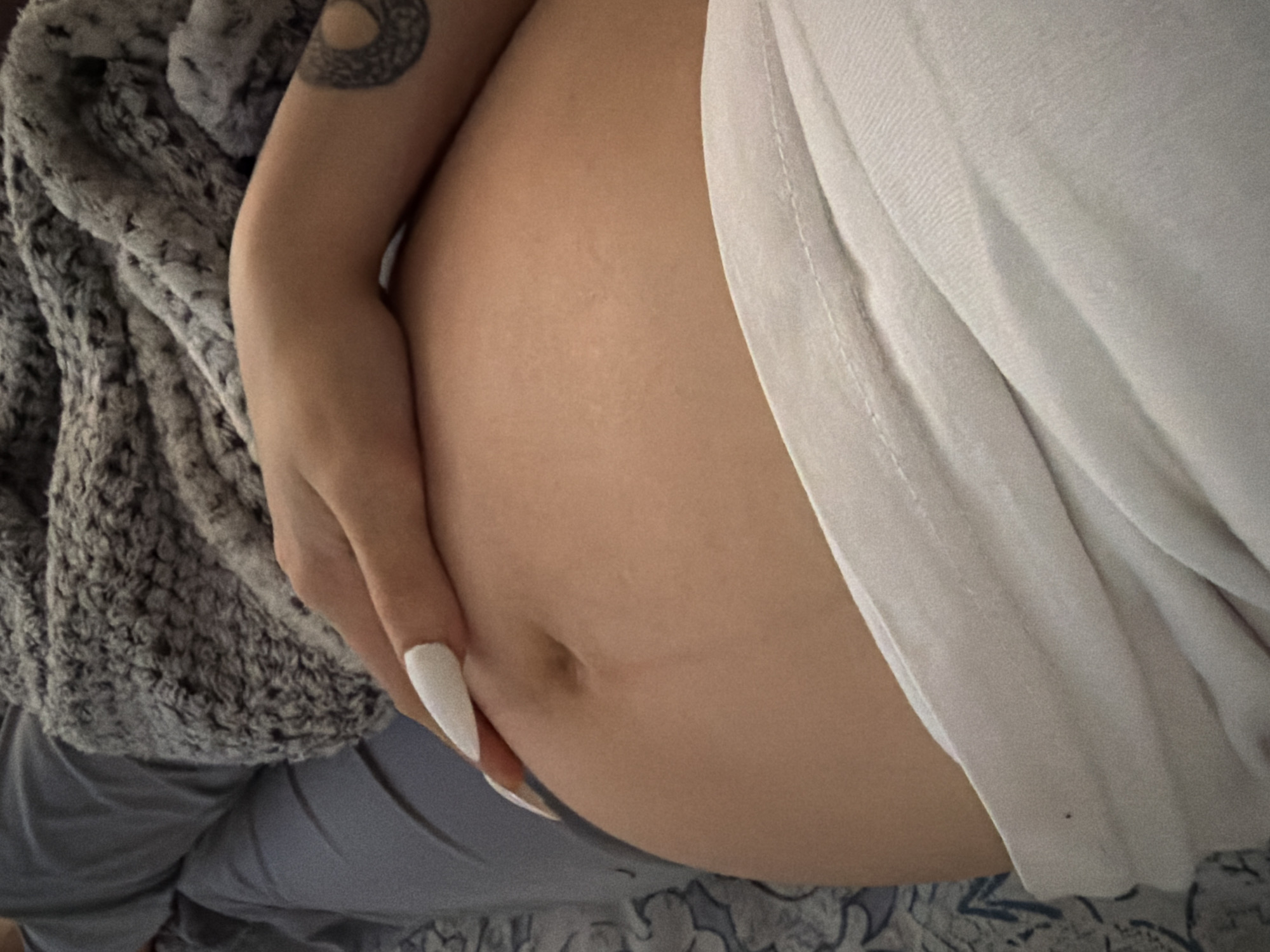 An individual's pregnant abdomen is visible, covered by a white garment and a grey, fuzzy blanket. The individual's right hand, with white nail polish, rests on their abdomen. A tattoo is visible on the individual's right arm. The background consists of grey bedding. There is no indication of the location being Tbilisi, Georgia.