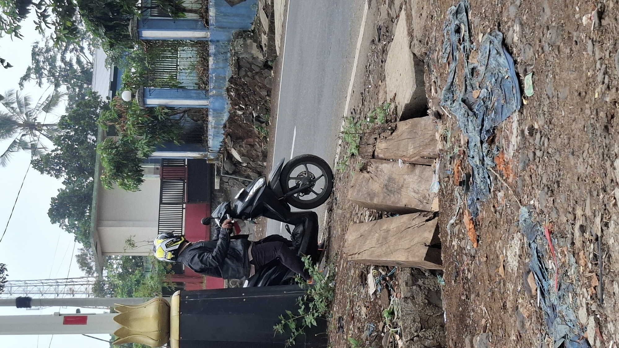 A person wearing a black leather jacket and a yellow and black helmet is sitting on a black motorcycle parked on the side of a road. The person's right hand is on the motorcycle's handlebar. The motorcycle has a silver rim on its front wheel. The scene is in Cilaku, Indonesia. The foreground shows a rough ground with debris, including pieces of wood and blue plastic sheeting. In the background, there is a road, a blue and white fence, a building, and some vegetation. A tall pole with red and white markings is visible to the left of the frame.