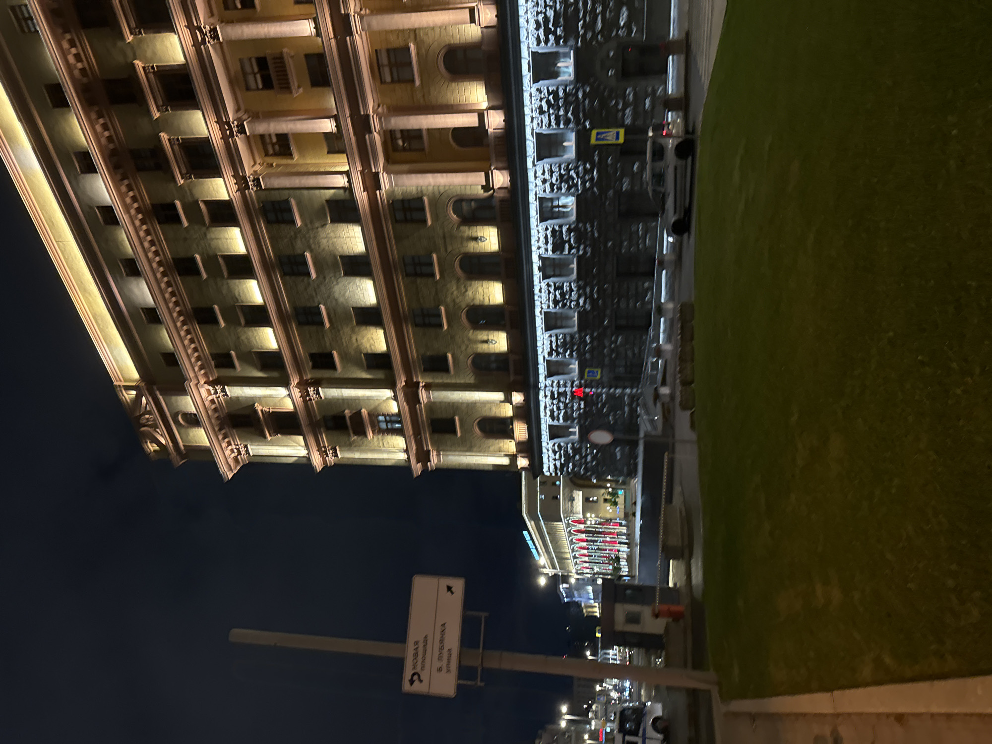 The image shows a nighttime view in Moscow, Russia, featuring architectural details. A tall building with lighted windows stands prominently on the left. To its right, there's a wall with stone-like texture, and a green lawn extends further to the right. In the lower part of the frame, a street sign with Russian text is visible. Further in the background, there are building lights and vehicles, indicating urban activity.