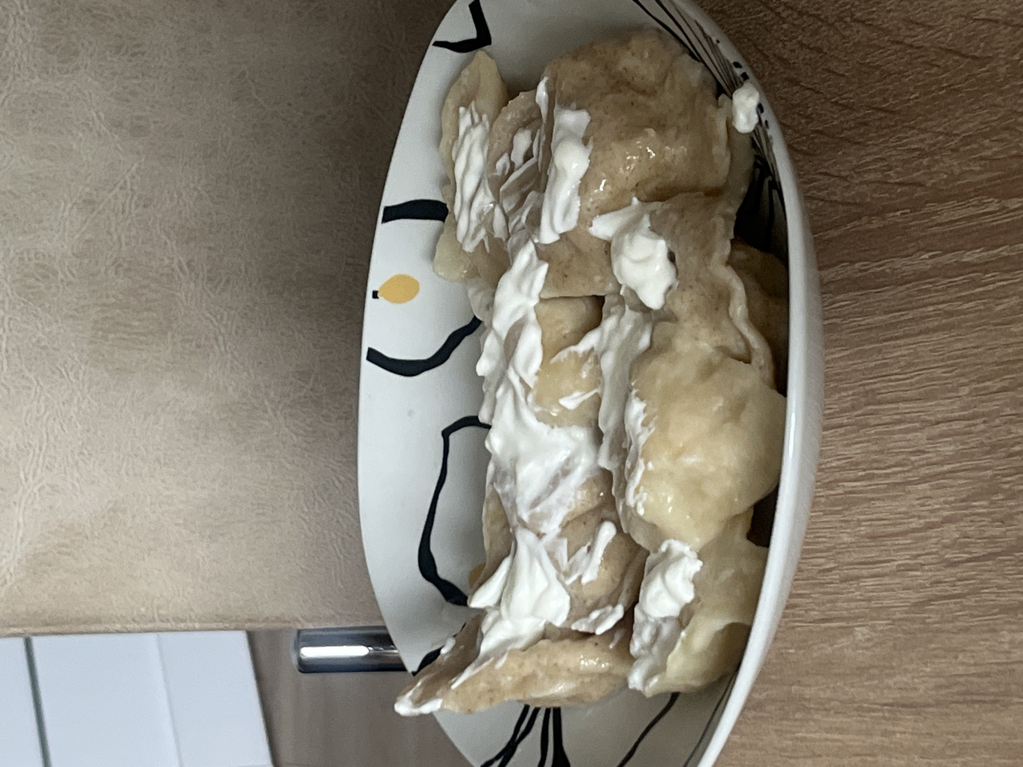 A close-up shot shows a bowl of dumplings topped with sour cream. The bowl has a white background with abstract black and yellow designs. The dumplings are arranged in the bowl, and the sour cream is dolloped on top of them. The bowl is placed on a wooden surface. In the background, a beige textured surface is visible, possibly a wall or upholstery. A silver object, resembling a part of a chair leg, is also visible to the left of the bowl. The scene appears to be an indoor setting. It is not possible to determine the location in the city of Rybnitsa, Moldova from the image.