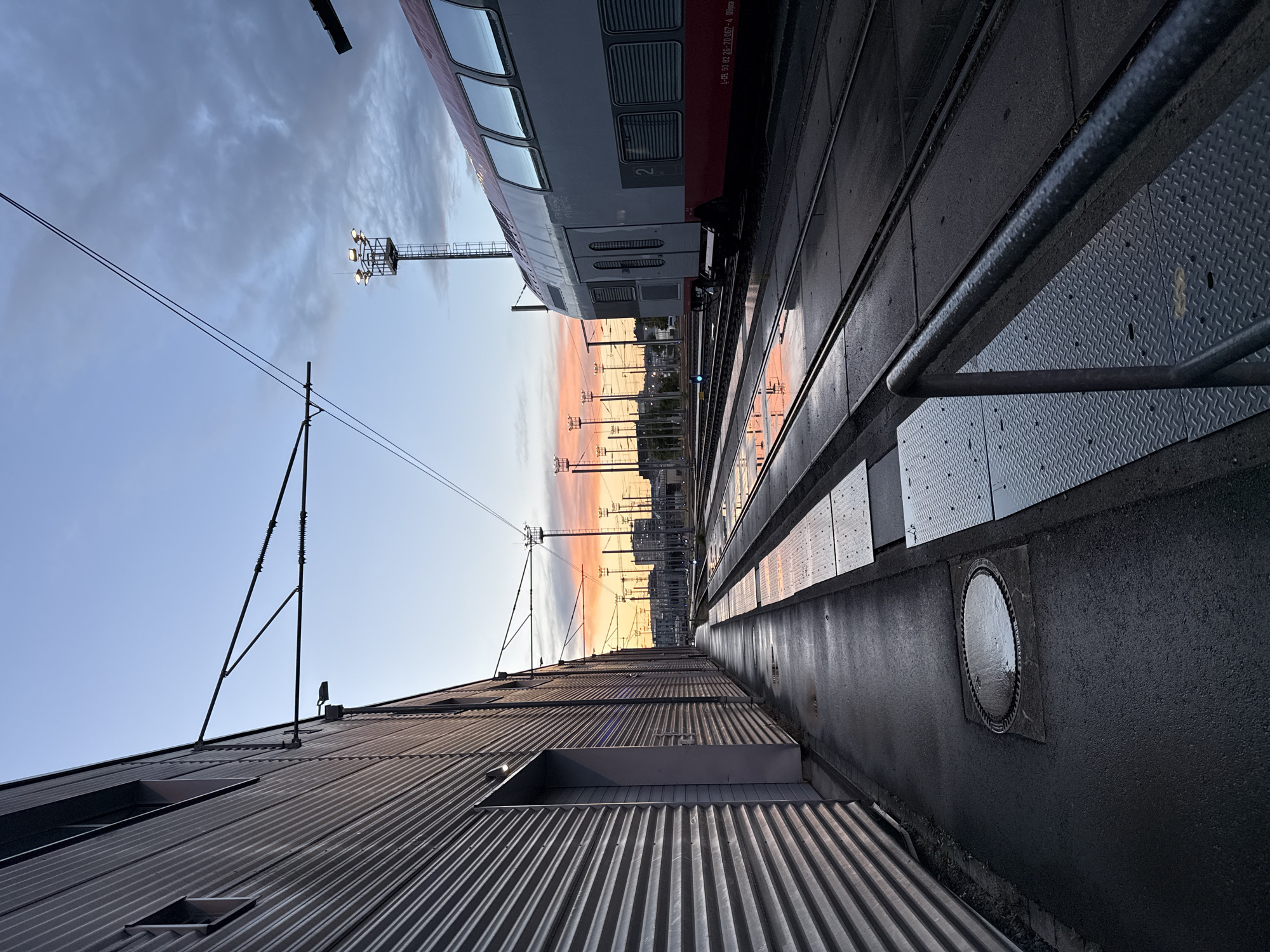 The image captures a serene railway scene during either sunrise or sunset, indicated by the striking gradient of warm orange, pink, and yellow light stretching across the horizon, blending into a soft blue sky with scattered clouds above. The perspective is low, looking down a set of railway tracks from a platform or adjacent walkway.
On the right side, a modern double-decker passenger train, predominantly grey with a red stripe, is stationary. The number "2" is visible on one of its doors, likely denoting a class designation. Along the train's side, text reads "1-B 50 82 26-70 067-4 Regio," suggesting it's a regional service, a common type of train operating in Luxembourg. The train's windows reflect the vibrant sky.
Multiple railway tracks extend into the background, accompanied by overhead power lines (catenary) and their support poles. A platform area runs alongside the tracks, featuring textured metal anti-slip plates and darker asphalt or concrete. A circular manhole cover is visible on this surface. Further back, a tall floodlight tower with multiple lamps illuminates the area.
On the left, a long, low-slung building with a corrugated metal roof and sides runs parallel to the tracks. Several skylights or roof windows are visible on its surface. The overall impression is one of quiet transition, with the industrial elements of the railway bathed in the atmospheric glow of the golden hour in Luxembourg.