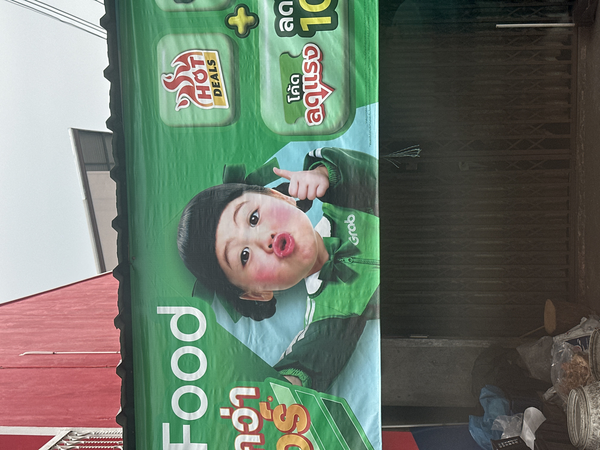 This media file captures a close-up of a vibrant green billboard advertising GrabFood. The billboard features a child dressed in a green Grab uniform with a playful expression, puckering their lips. The text on the billboard is in Thai and English, promoting "HOT DEALS" and offering a discount of "10" with a code labeled "ลดแรง" (which translates to "strong discount"). The "Food" text is prominently displayed in large white lettering. The setting appears to be outdoors, with a hint of overcast sky in the background and a red building visible to the left, suggesting an urban environment. The overall impression is one of a promotional advertisement for a food delivery service, likely aimed at families or younger audiences due to the child's imagery.
