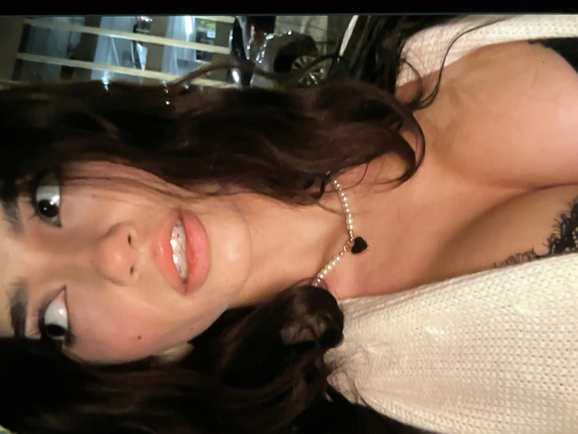 A young woman with long, dark, wavy hair is captured in a close-up, self-taken photograph. She is looking to her left with her mouth slightly open, revealing some teeth. Her lips are glossy and her eyebrows are dark and defined. She is wearing a white knit cardigan over a black lace top, and a pearl necklace with a black heart pendant rests on her décolletage. The background appears to be an urban environment at night, with blurry lights and what seem to be the exterior of a building with vertical elements and a parked vehicle. The lighting is dim and appears to be artificial.