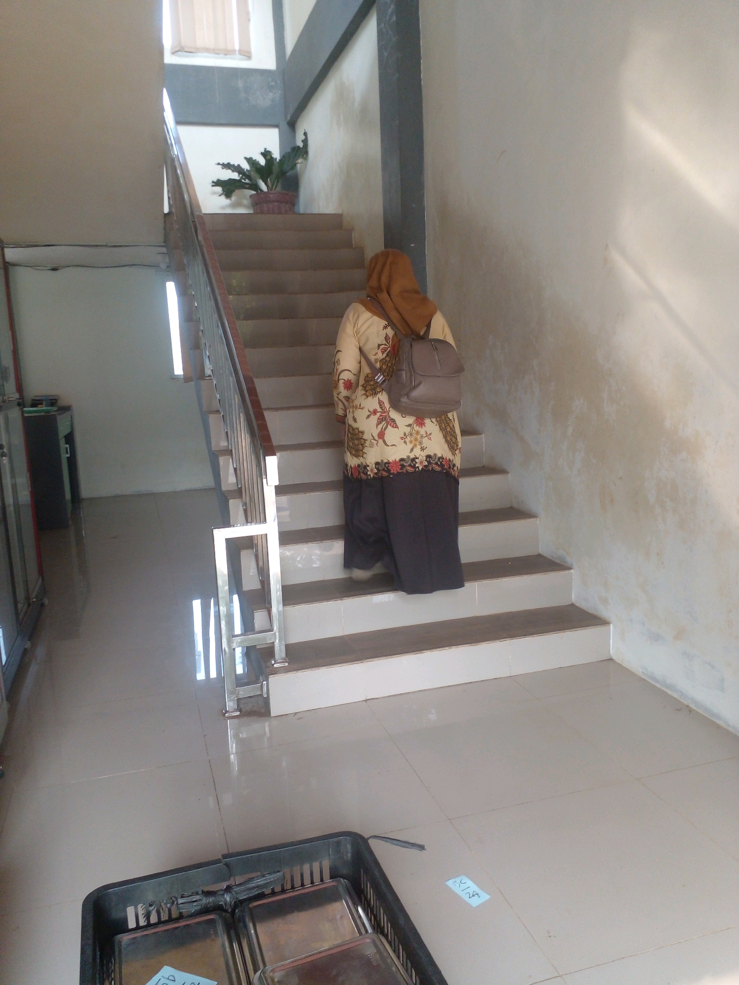 A woman wearing a tan hijab and a patterned tunic walks up a light-colored concrete staircase. She has a grey backpack slung over her right shoulder. The staircase has a metal railing with a wooden top. To the left of the staircase is a white wall, and to the right is another light-colored wall. A potted plant sits on the landing at the top of the first flight of stairs. Below the staircase, on a tiled floor, is a black plastic basket containing metal trays. The scene appears to be in a building, possibly an office or school, located in Banyuputih, Indonesia.