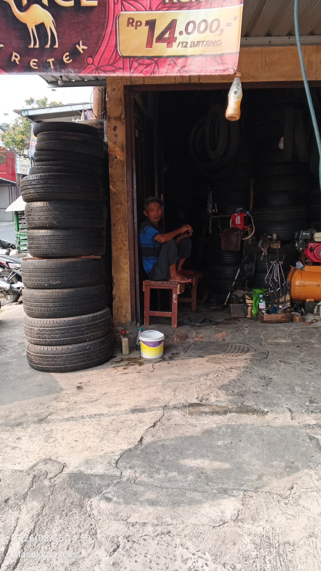 A man is sitting on a low stool in front of a tire shop located in Malang, Indonesia. The shop is open to the street, revealing stacks of tires both inside and outside. The man is wearing a blue and black striped shirt and dark pants, with a gray cap on his head. He is barefoot and looking towards the camera. Behind him, inside the shop, are more tires, tools, and a compressor. To the left of the shop entrance, a stack of tires is piled high on the concrete ground. A sign above the shop displays "RP 14.000,- /12 BATANG" and an image of a camel. The ground in front of the shop is cracked and uneven concrete.