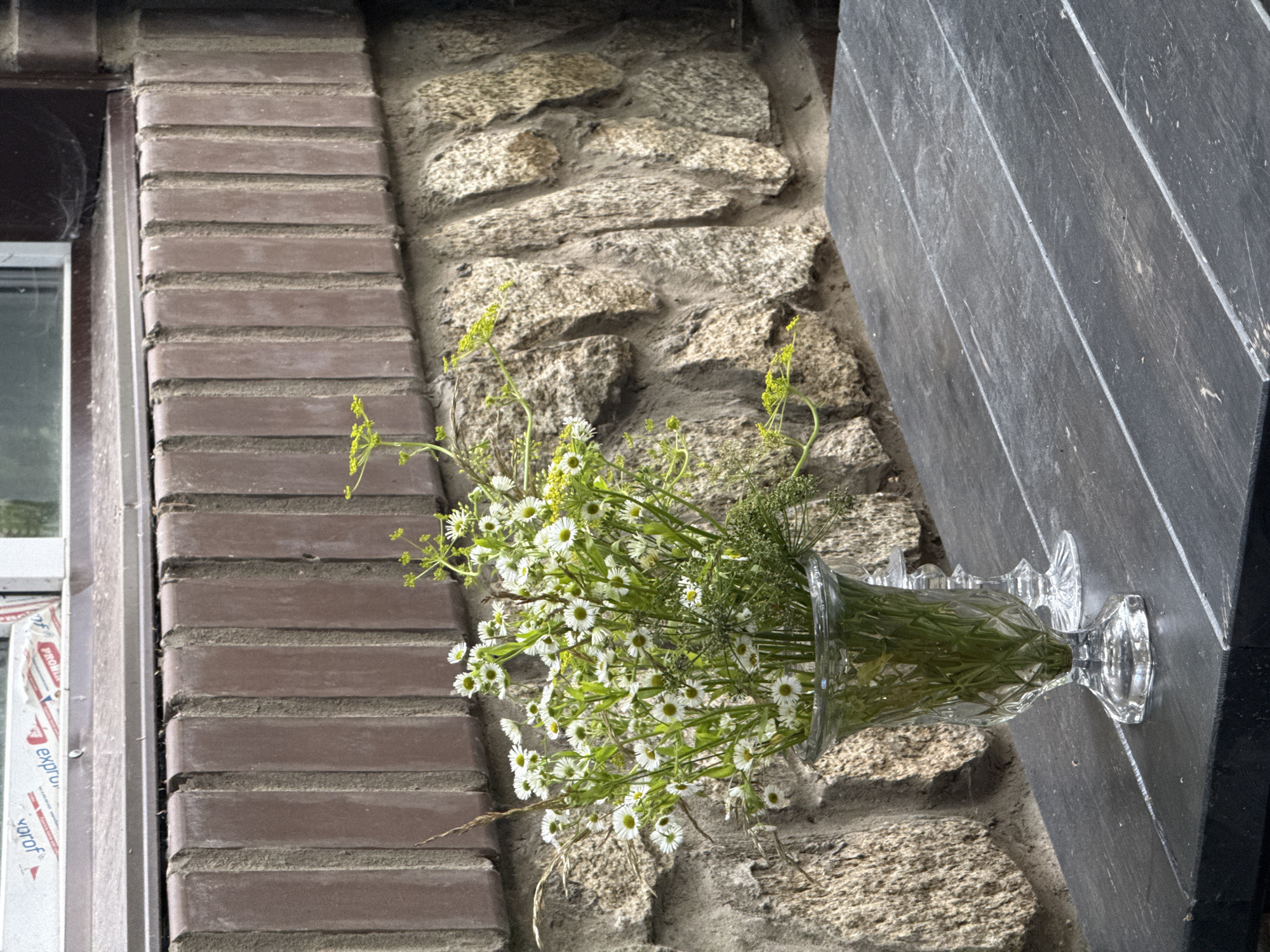 A bouquet of small white wildflowers with yellow centers and green foliage is displayed in a clear glass vase with a patterned surface. The vase is placed on a dark-colored wooden table. In the background, a textured stone wall is visible above a row of brown bricks that appear to form a decorative border. A portion of a window and a white framed structure are visible at the top of the image. The setting appears to be outdoors, possibly a patio or garden area. There are no people or animals present. The scene is static and devoid of any action. The image does not provide information that would allow for the identification of the city of Perm, Russia, or any specific location within it.
